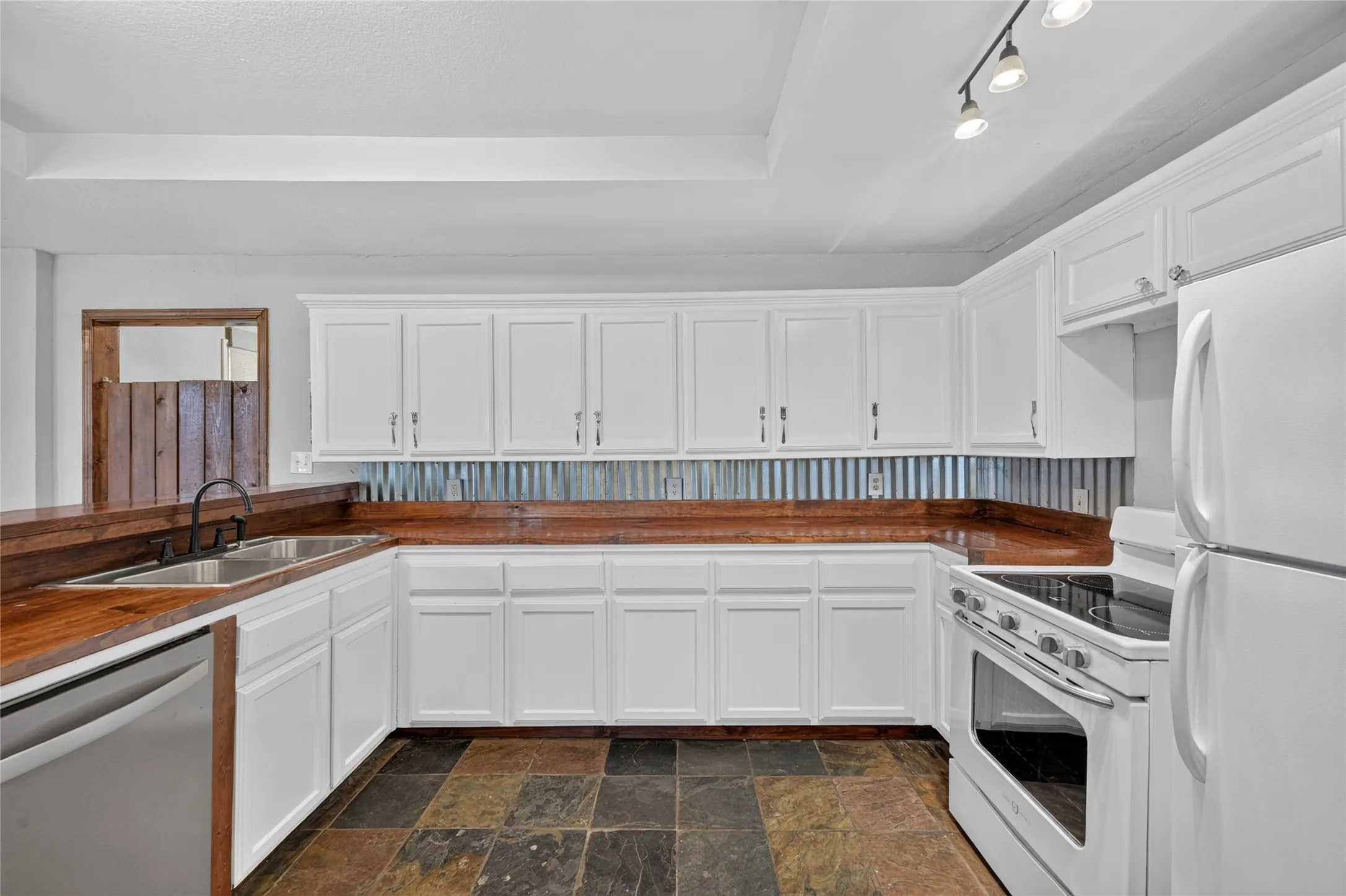 White cabinetry with wood-finish countertops, corrugated metal backsplash, track lighting, and multi-toned tile flooring