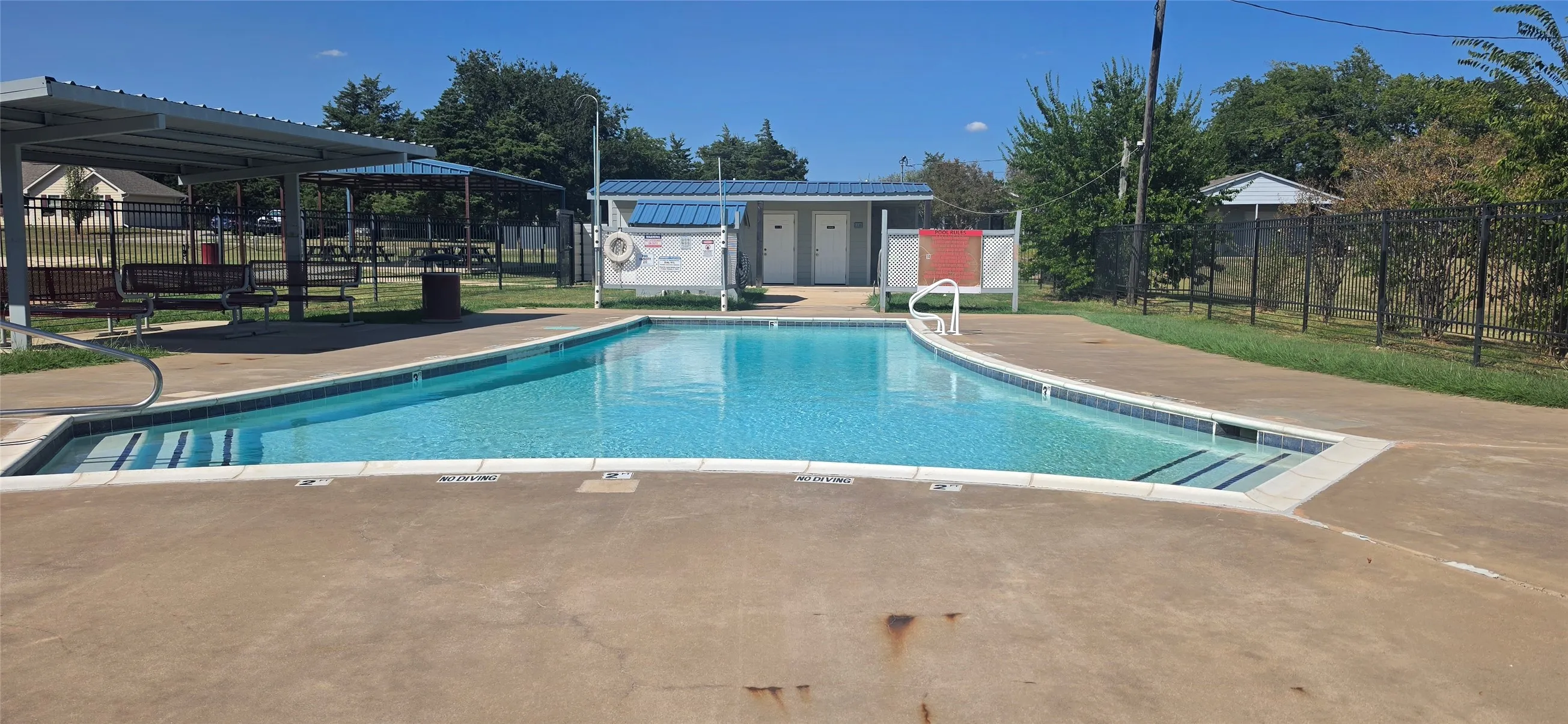 Outdoor swimming pool with integrated steps, surrounding concrete decking, metal handrails, and a nearby covered seating area