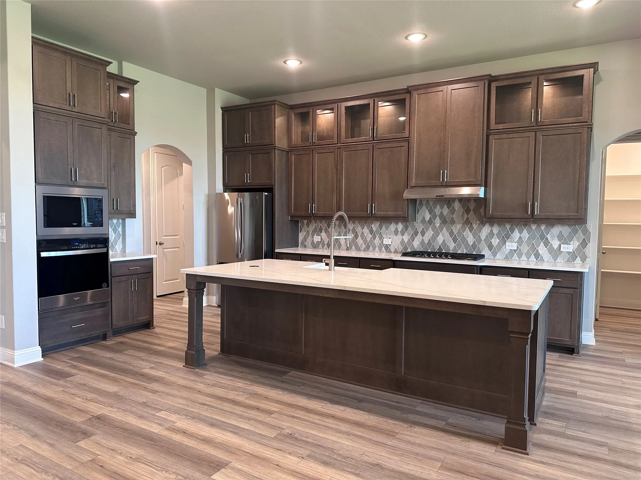 Kitchen featuring a large central island with a light-toned countertop, extensive cabinetry with integrated lighting, stainless steel appliances, a geometric-patterned backsplash, and wood-finish flooring
