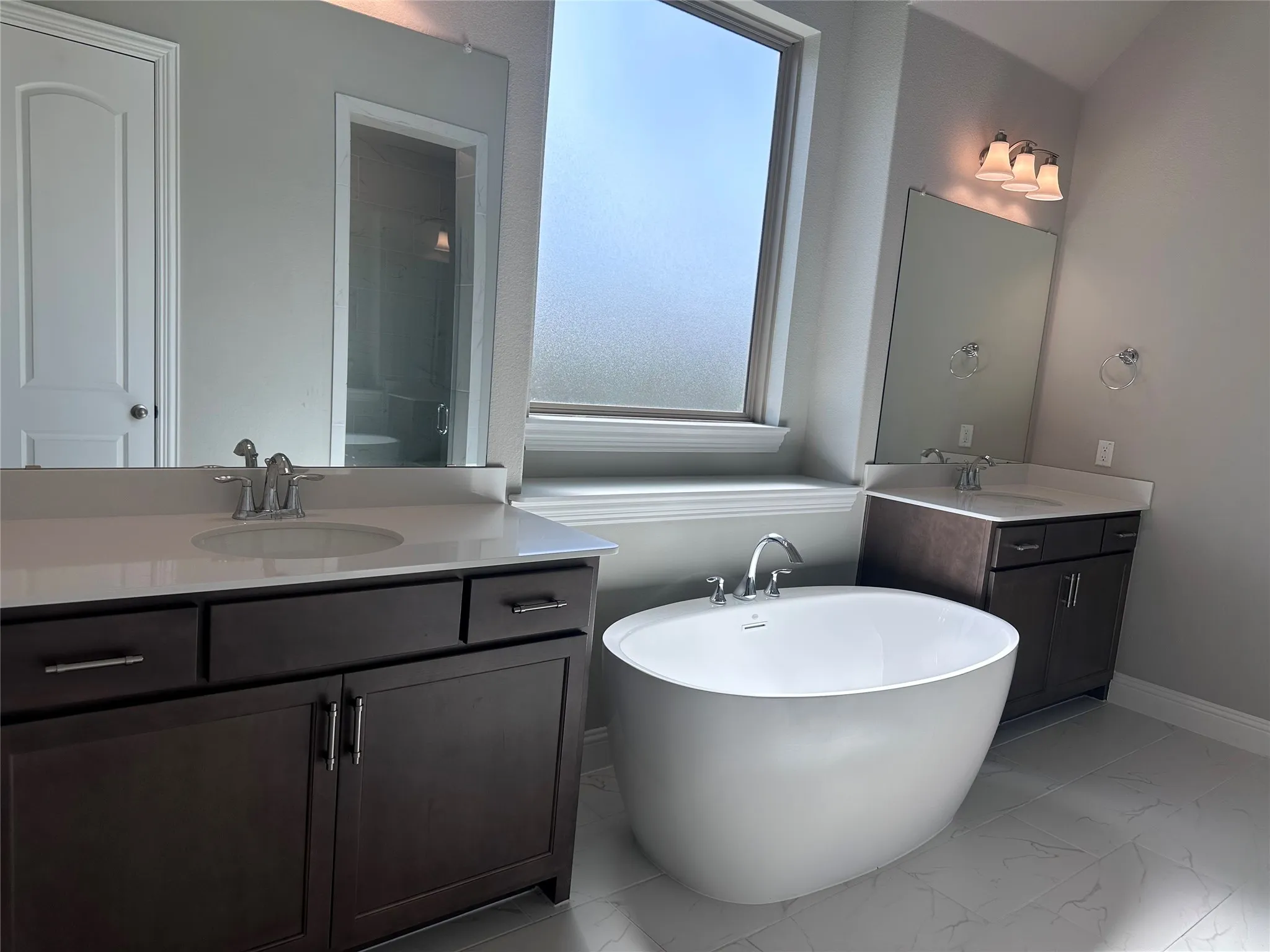 Dual vanities with white countertops, freestanding soaking tub, large frosted window, and light gray tiled flooring