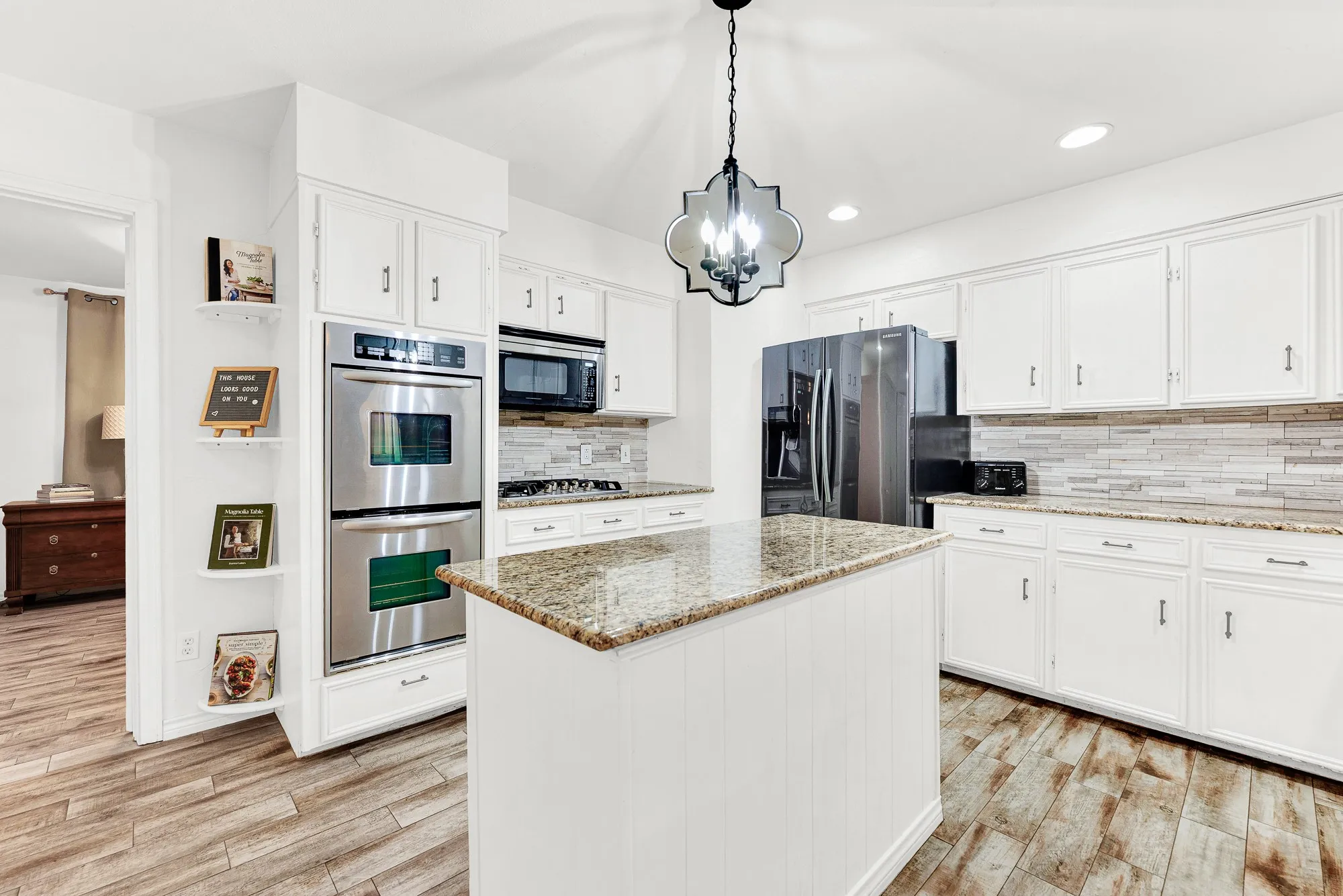This angle emphasizes the built-in double ovens, cooktop area, and warm granite countertops beneath a statement pendant light. Imagine baking weekend treats while keeping your favorite cookbooks organized on the built-in corner display shelves.