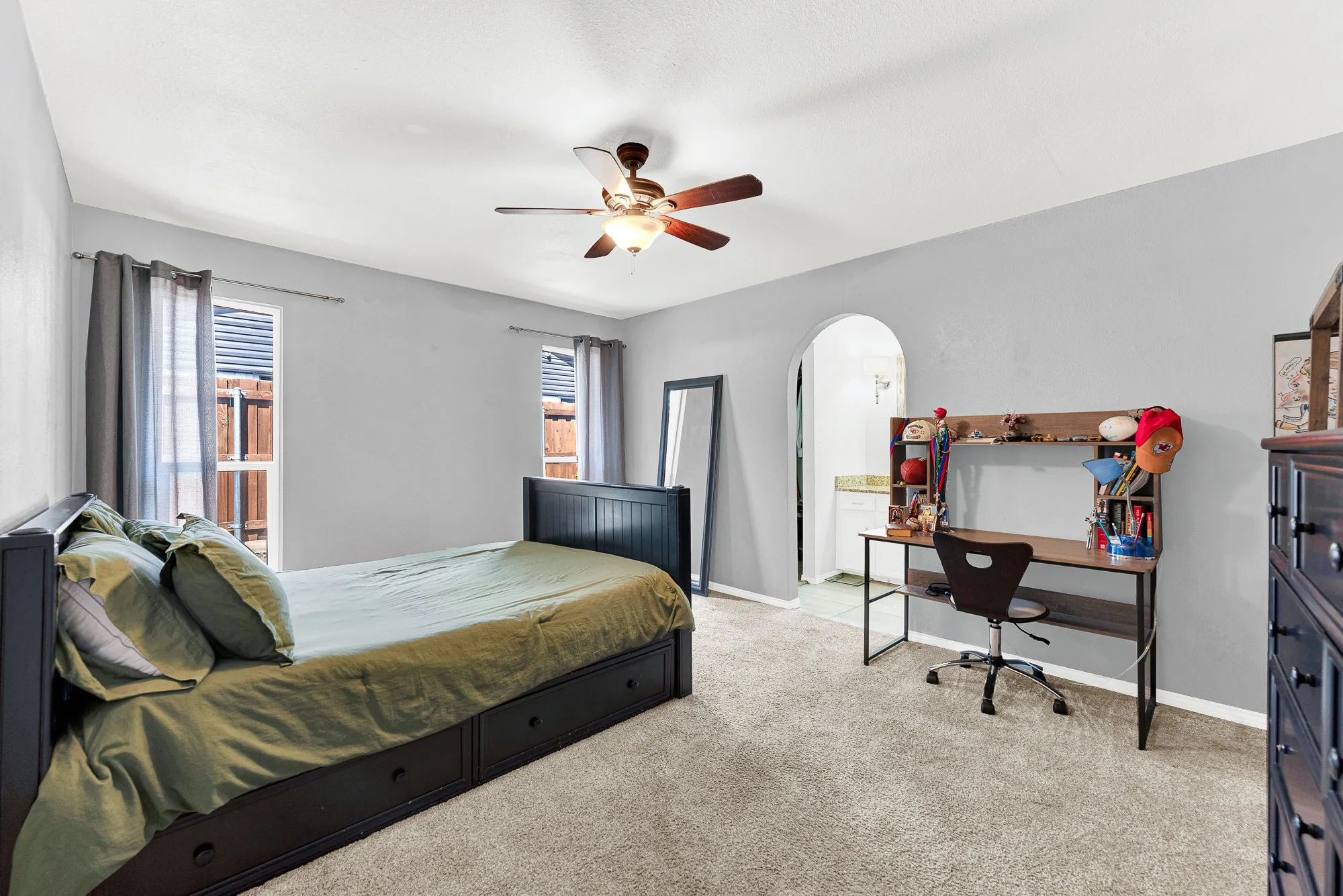 An arched opening frames a private ensuite bath, adding tailored architectural detail to this light-filled secondary bedroom. Soft carpet, neutrally painted walls, and a ceiling fan create a comfortable and functional retreat.