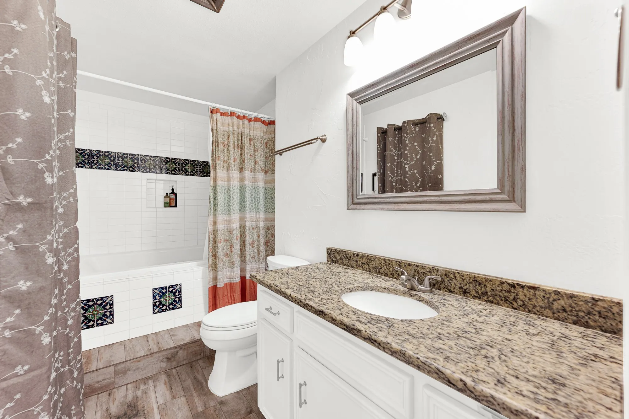 The primary en-suite bathroom boasts a spacious granite vanity, framed accent mirror, and detailed decorative tile inlays surrounding the shower-tub combo. Refreshing morning routines feel elevated in this private, clean sanctuary.