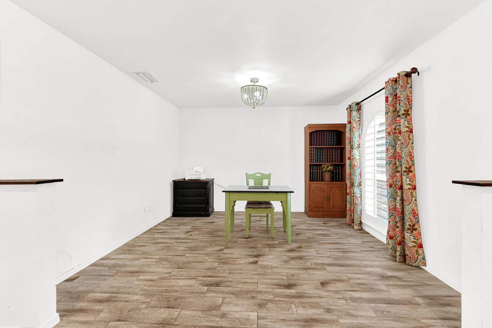 Recessed lighting and a beaded chandelier illuminate a dedicated work nook at the far end of the open flex room. Clean white walls and warm flooring provide a crisp, blank canvas ready to accommodate executive desks, crafting tables, or cozy reading chairs.
