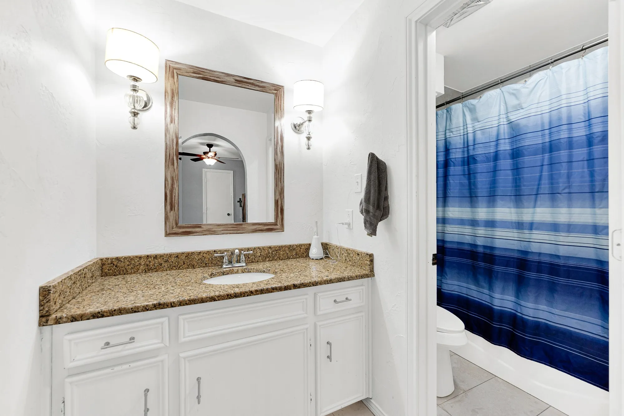 Inside the ensuite bath, a granite vanity with crisp white cabinetry offers ample daily storage. A wood-framed mirror flanked by classic sconces pairs nicely with the separate wet area, which features clean white tile and an easy-care tub-shower combinatio