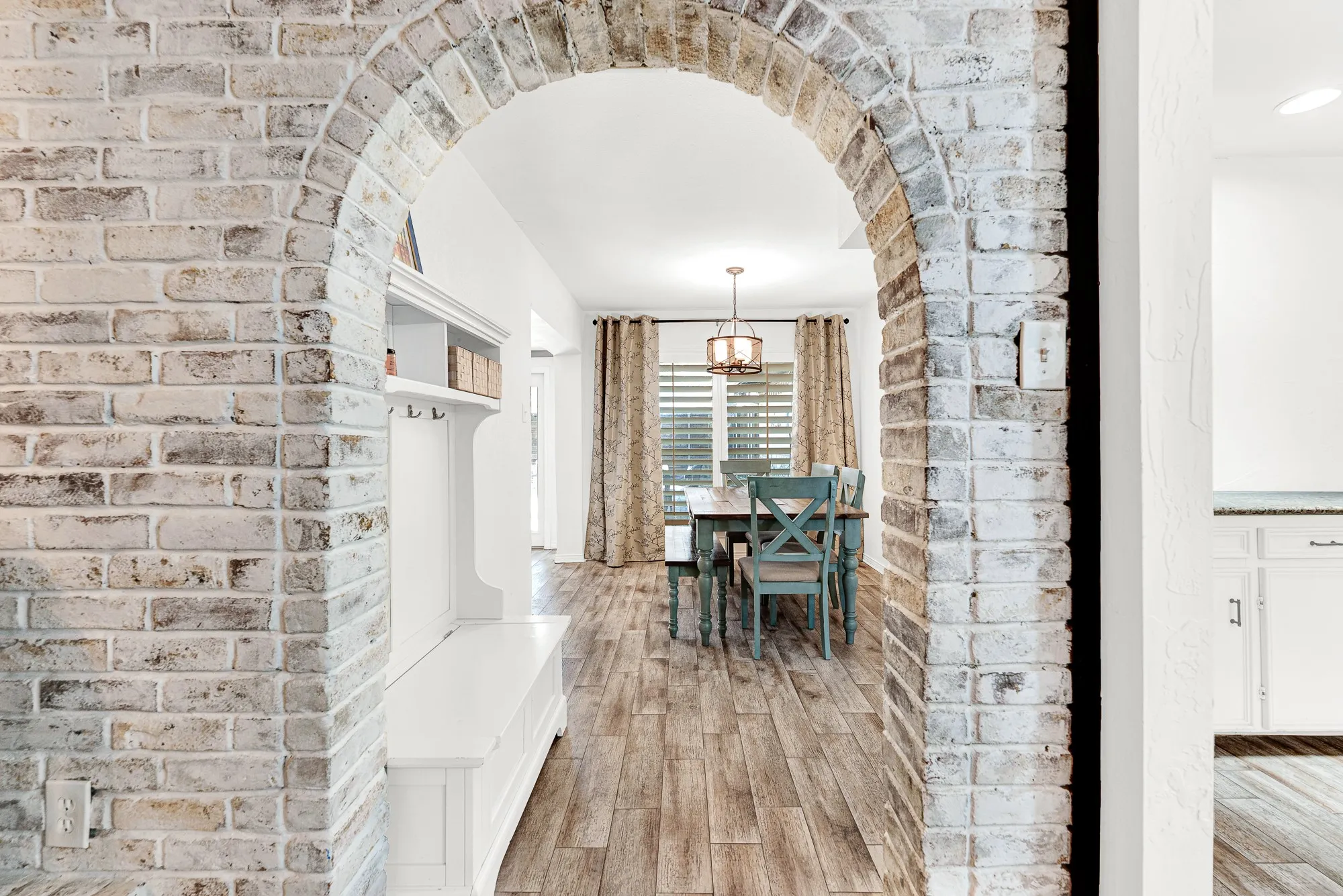 A whitewashed brick archway frames the casual dining space, featuring warm wood-look floors, large windows with natural light, and a charming pendant fixture. It offers an inviting setting for sharing weeknight family dinners or catching up over morning coffee
