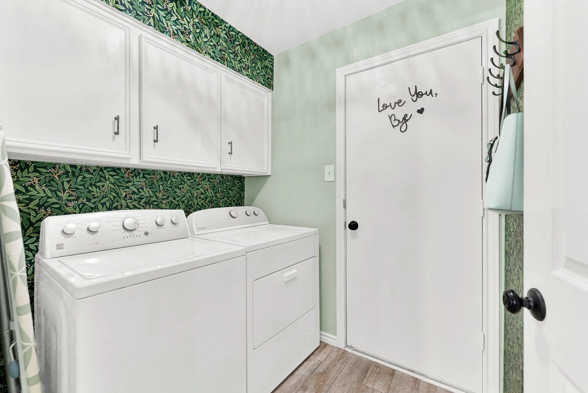 Stylish botanical wallpaper and soft green walls bring fresh charm into the dedicated utility room. Crisp white storage cabinets above the washer and dryer space keep laundry supplies organized right along the direct path to the garage.