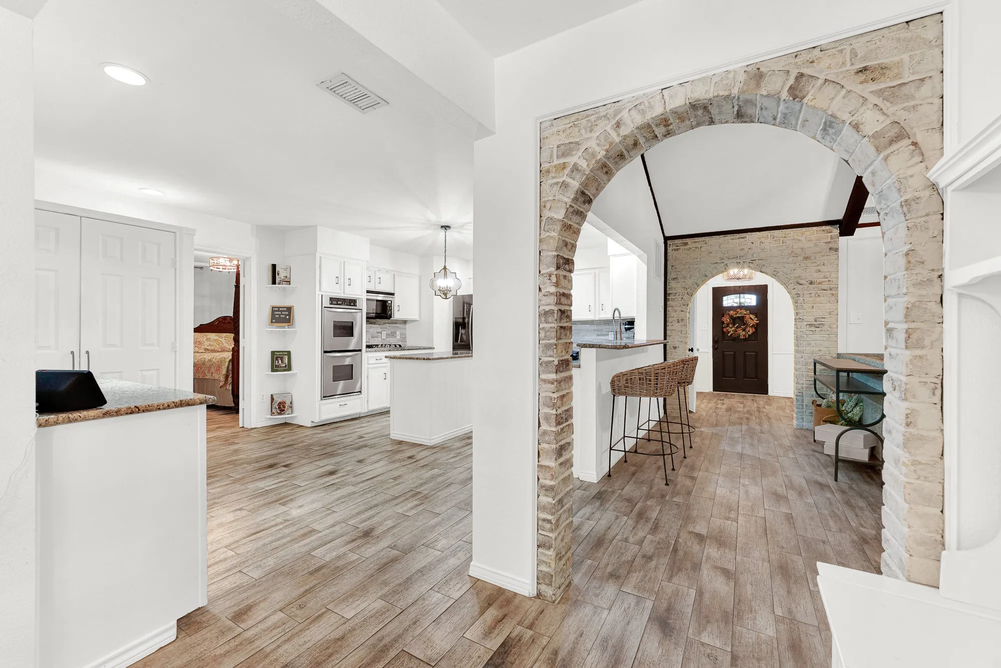 A striking whitewashed brick archway anchors the main level, framing open sightlines across the wood-look tile floors. This perspective connects the kitchen layout, central dining nook, and entry foyer in one seamless flow.