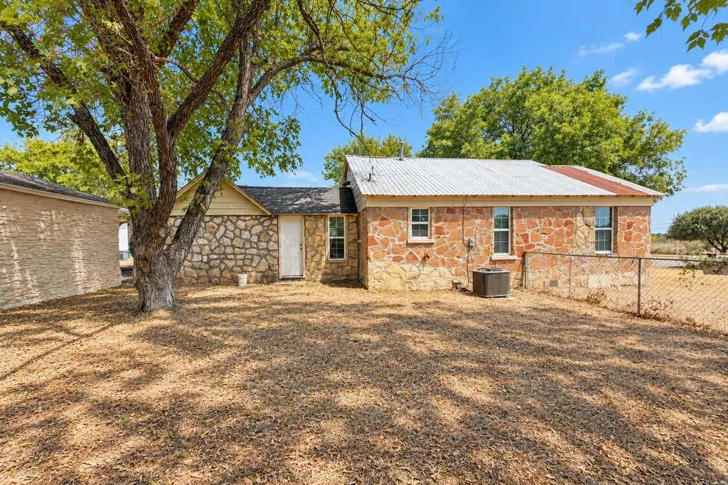 Side yard beside the stone main home with a large shade tree and chain-link fencing.