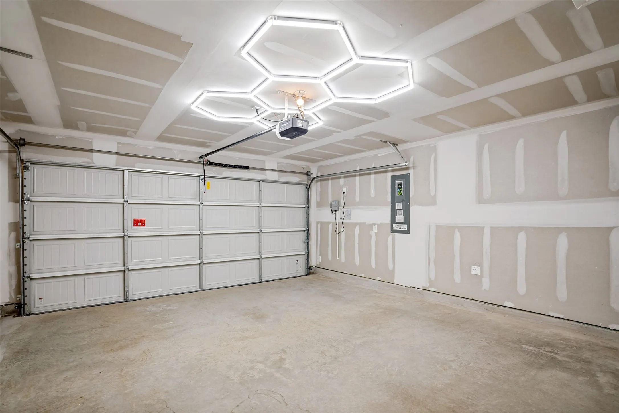 Concrete flooring, automatic garage door opener, modern hexagonal ceiling light fixture, exposed drywall, and electrical panel