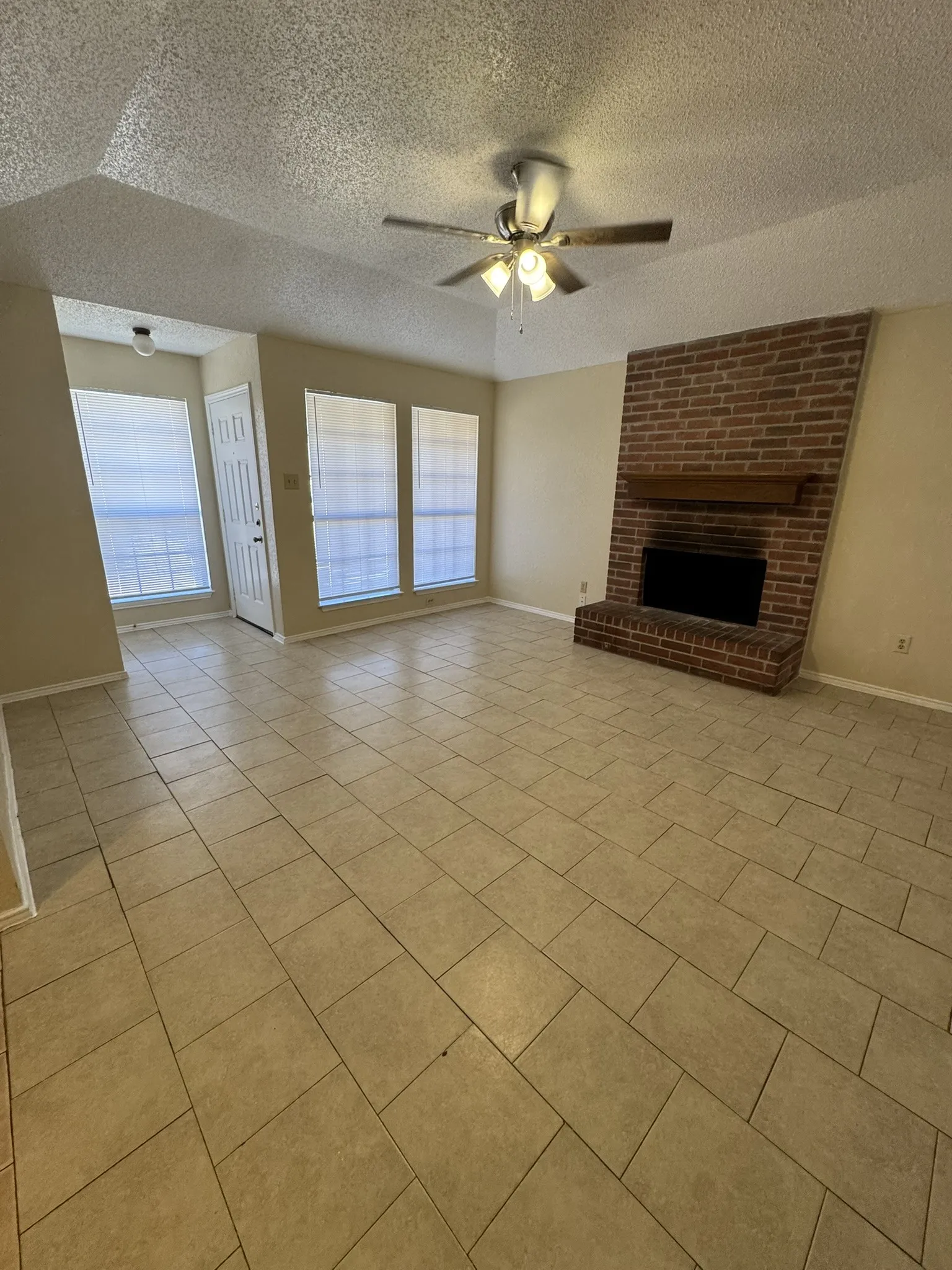 Spacious living area featuring tile flooring, a prominent brick fireplace with a wood mantle, multiple windows, and a ceiling fan