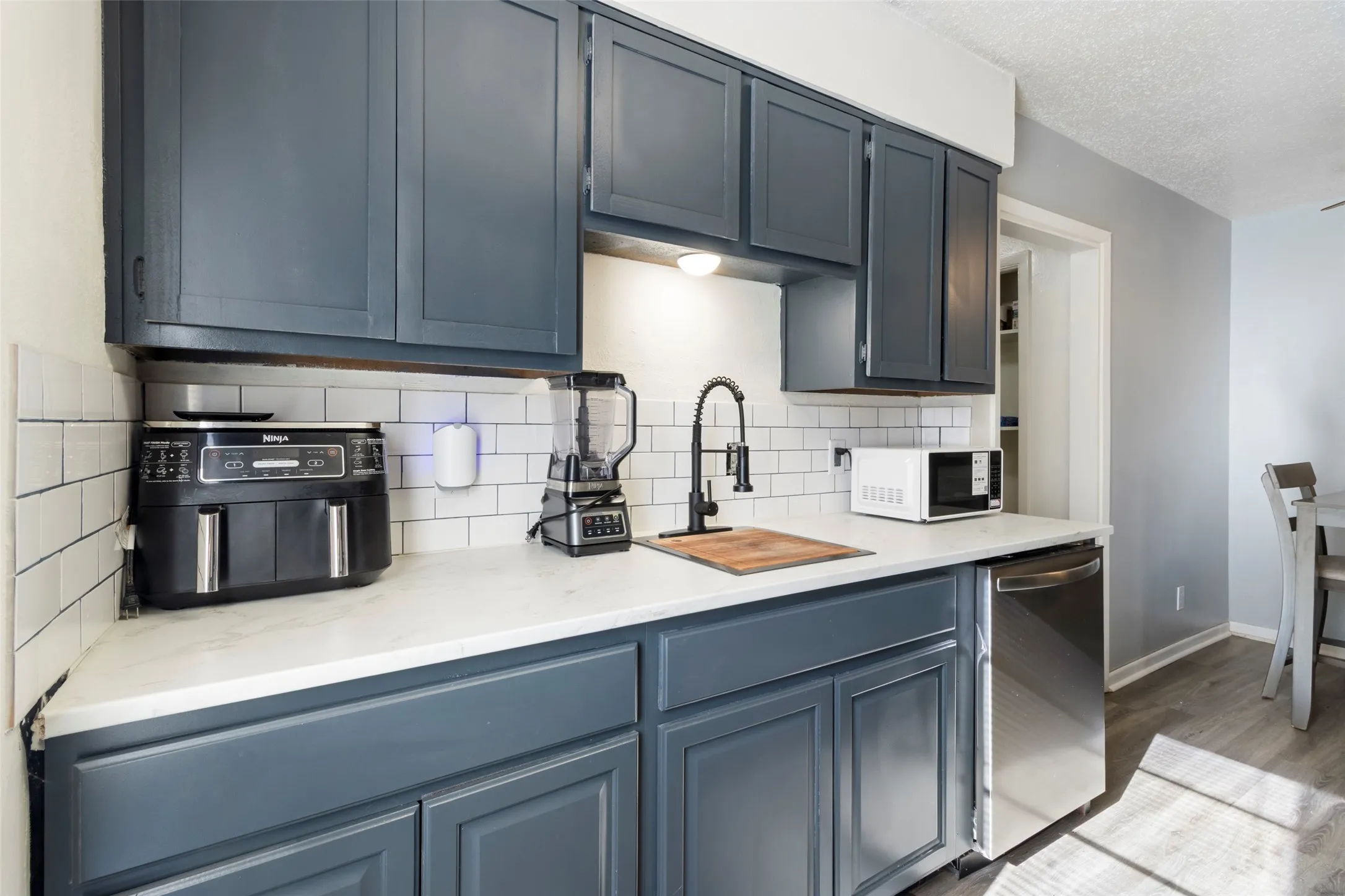Dark-toned cabinetry, white subway tile backsplash, stainless steel dishwasher, white countertops, and wood-finish flooring
