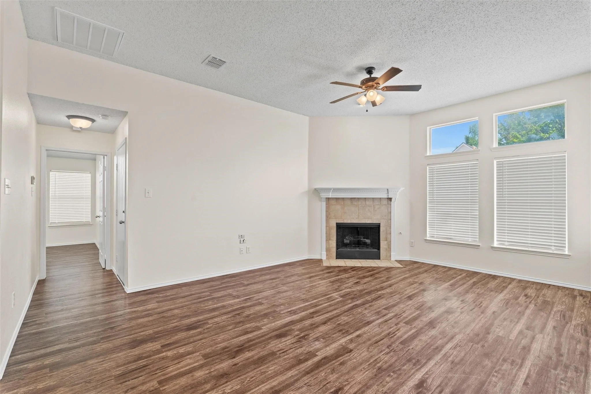Spacious living area featuring wood-finish flooring, a tiled fireplace with mantel, multiple windows for natural light, and a ceiling fan