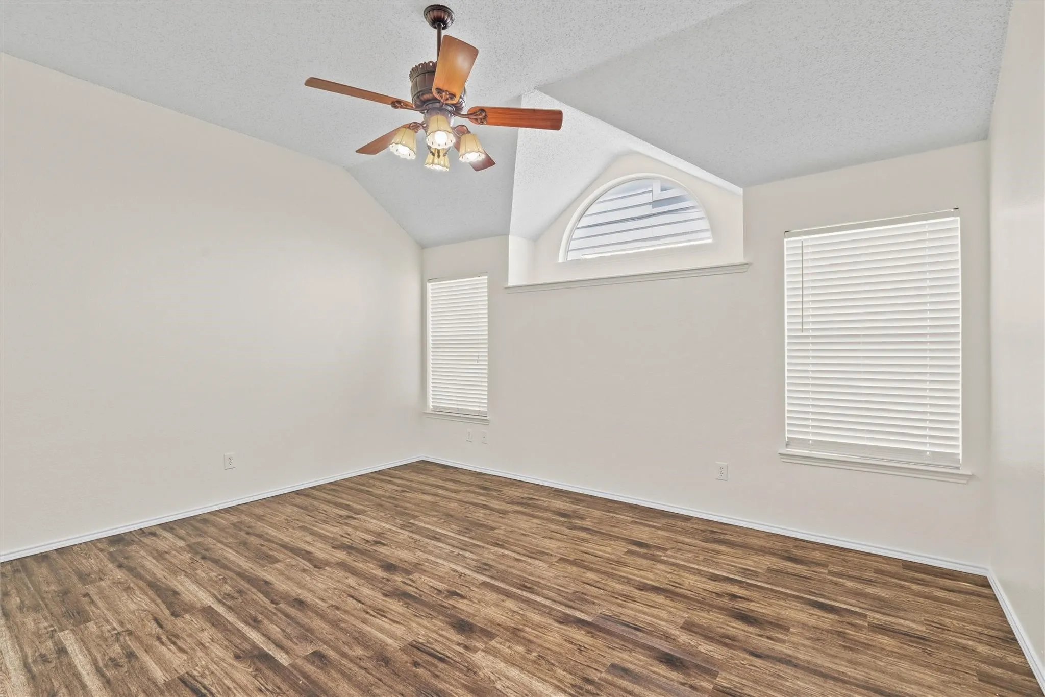 Vaulted ceiling featuring a ceiling fan with light kit, wood-finish flooring, and multiple windows including an arched transom window