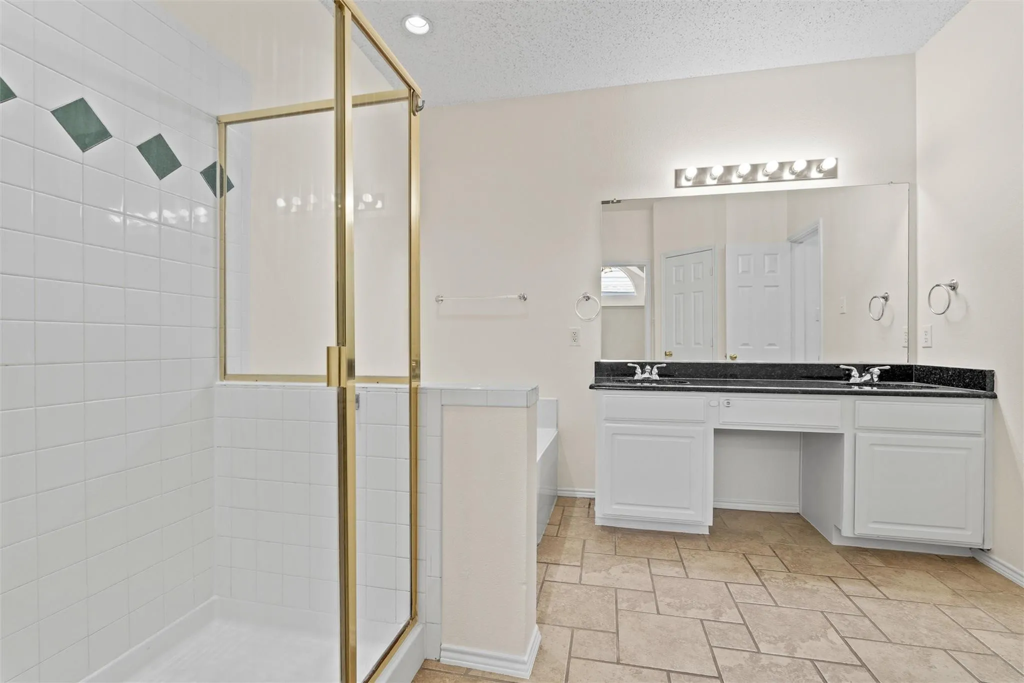 Walk-in shower with gold-tone frame, white subway tile surround with accent tiles, double vanity with dark countertop, large vanity mirror, and tile flooring