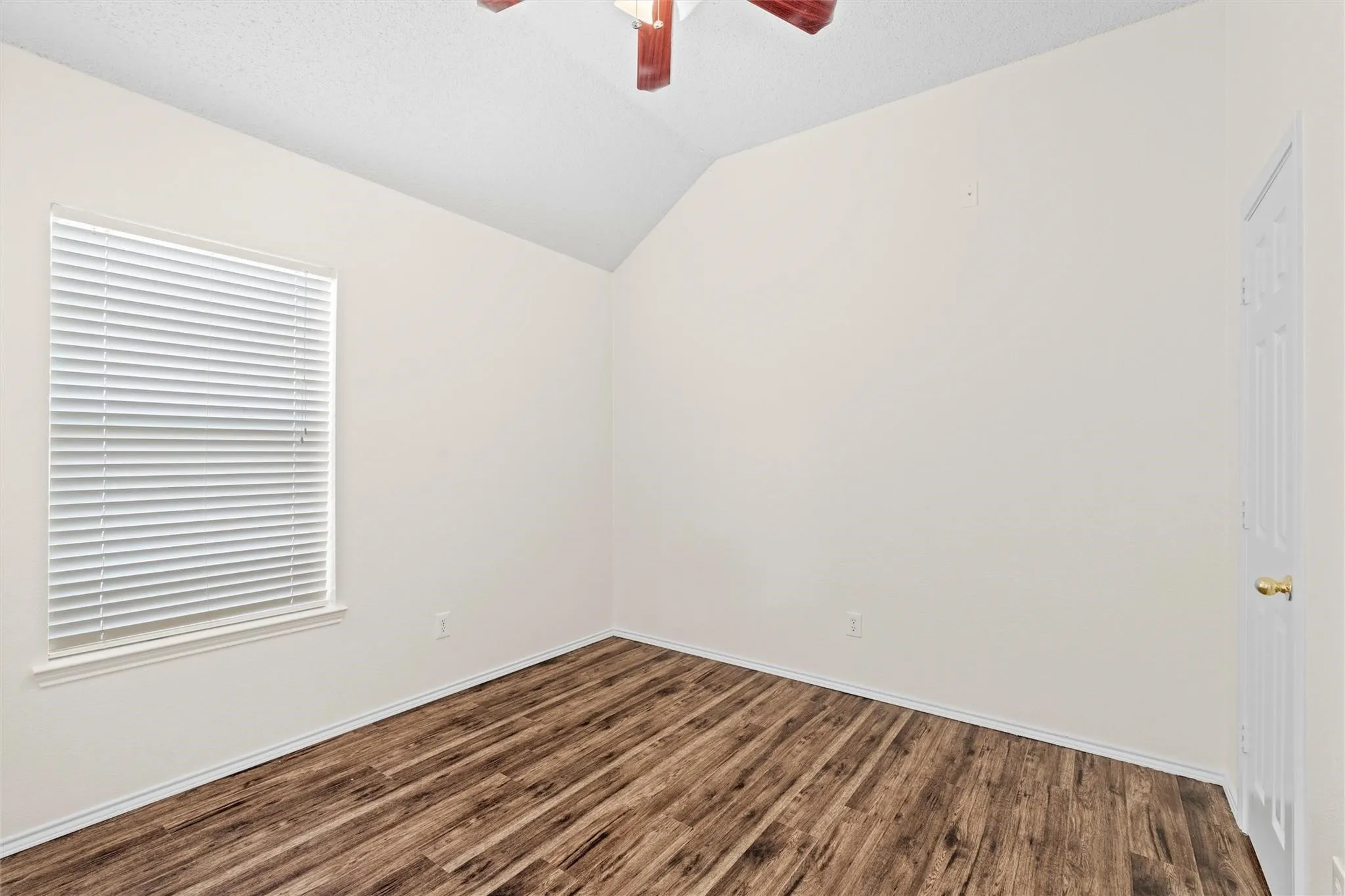 Sloped ceiling room featuring wood-finish flooring, a window with horizontal blinds, and a ceiling fan