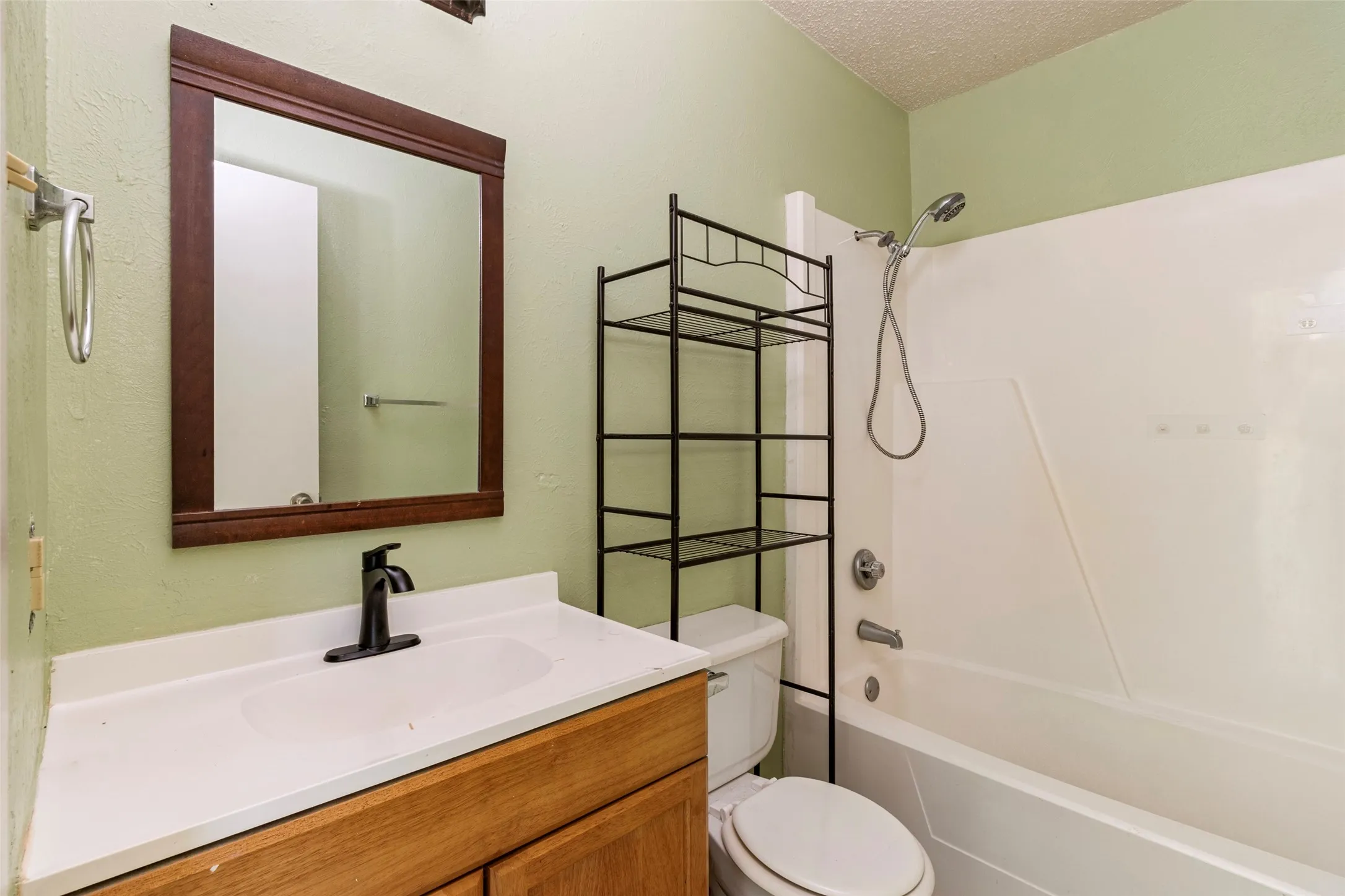 Wood-finish vanity with a white countertop and integrated sink, framed mirror, matte black faucet, and a combination shower-tub