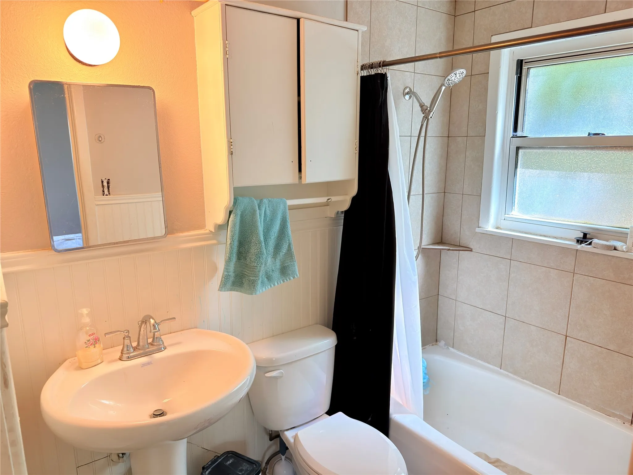 Pedestal sink with chrome fixtures, wall-mounted medicine cabinet, beadboard wainscoting, tiled bath enclosure with built-in shelving, and a window for natural light