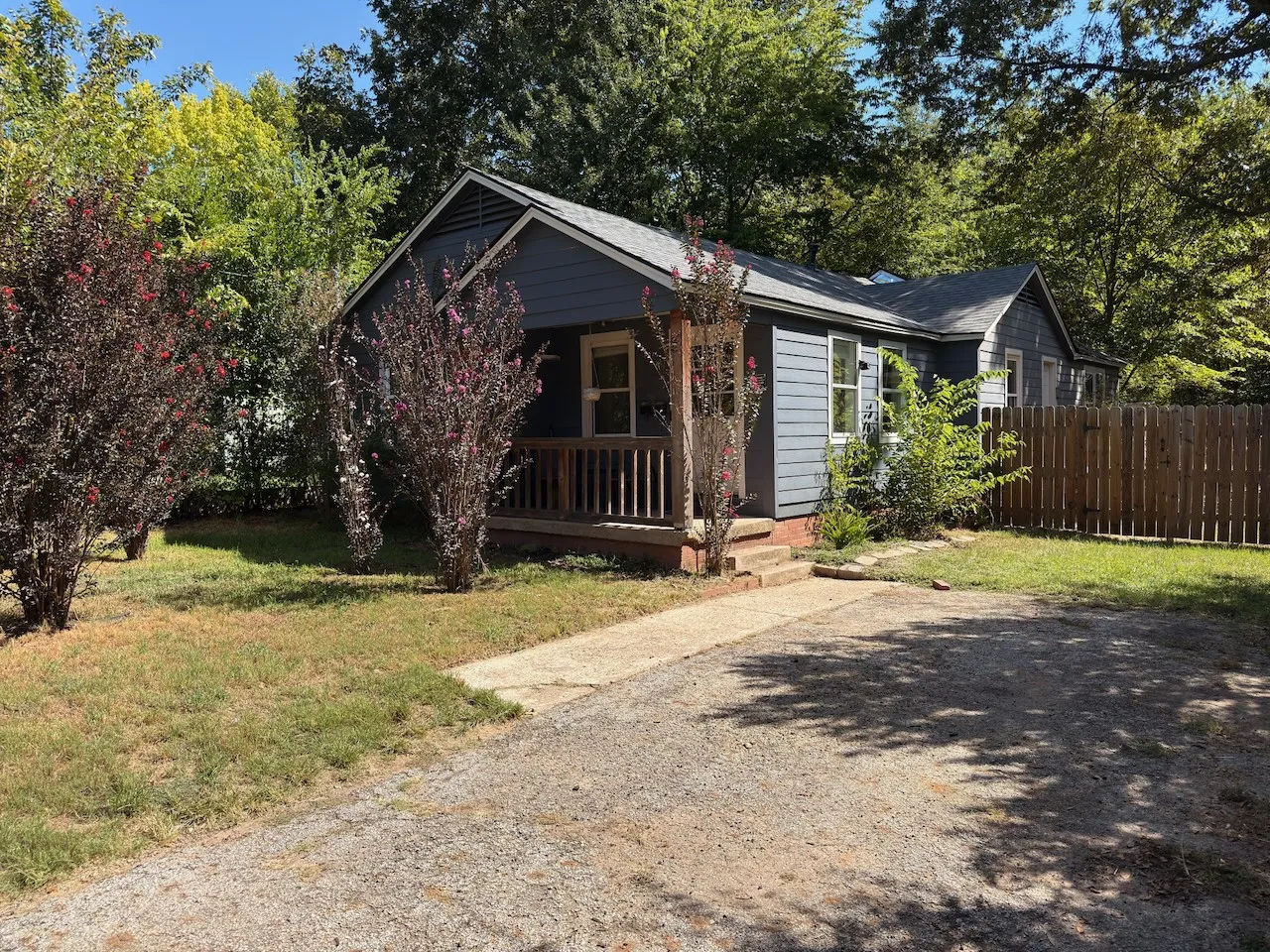 Blue exterior siding, wood fence, front porch with wood railings, gravel driveway, and mature landscaping