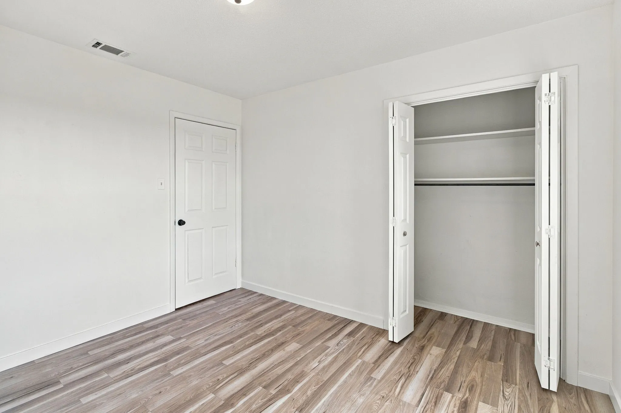 LVP flooring, white walls, white paneled door with black hardware, and a built-in closet with bifold doors and shelving