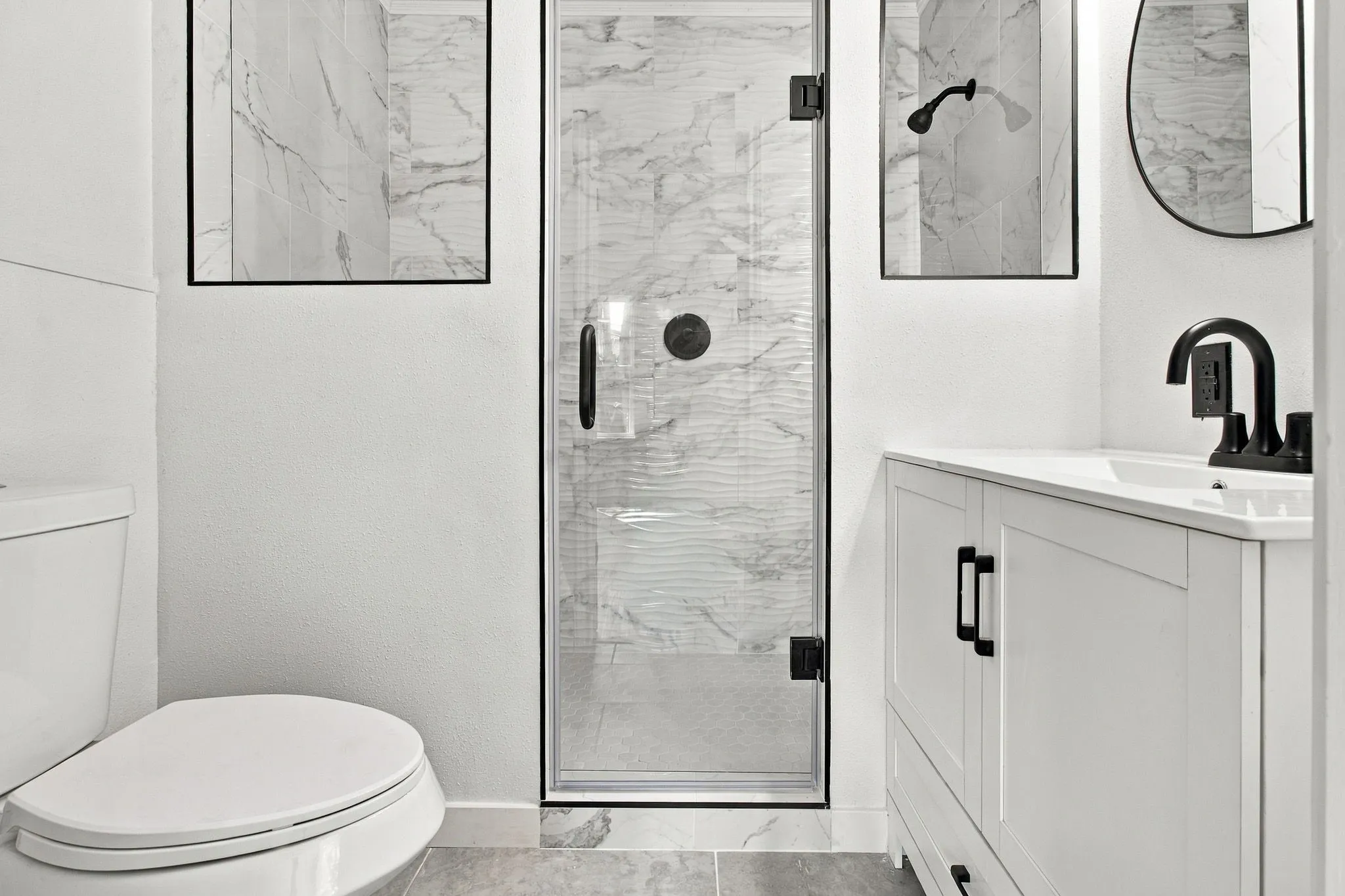 Bathroom featuring a glass-enclosed shower with marble-look wall tile, a single vanity with shaker-style cabinetry, and matte black fixtures