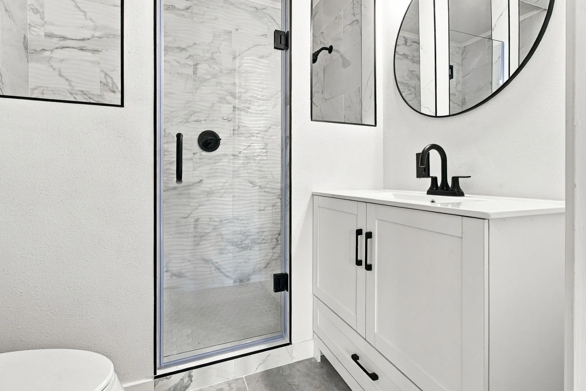 Walk-in shower with glass enclosure, single vanity with white countertop, matte black fixtures, and a frameless oval mirror