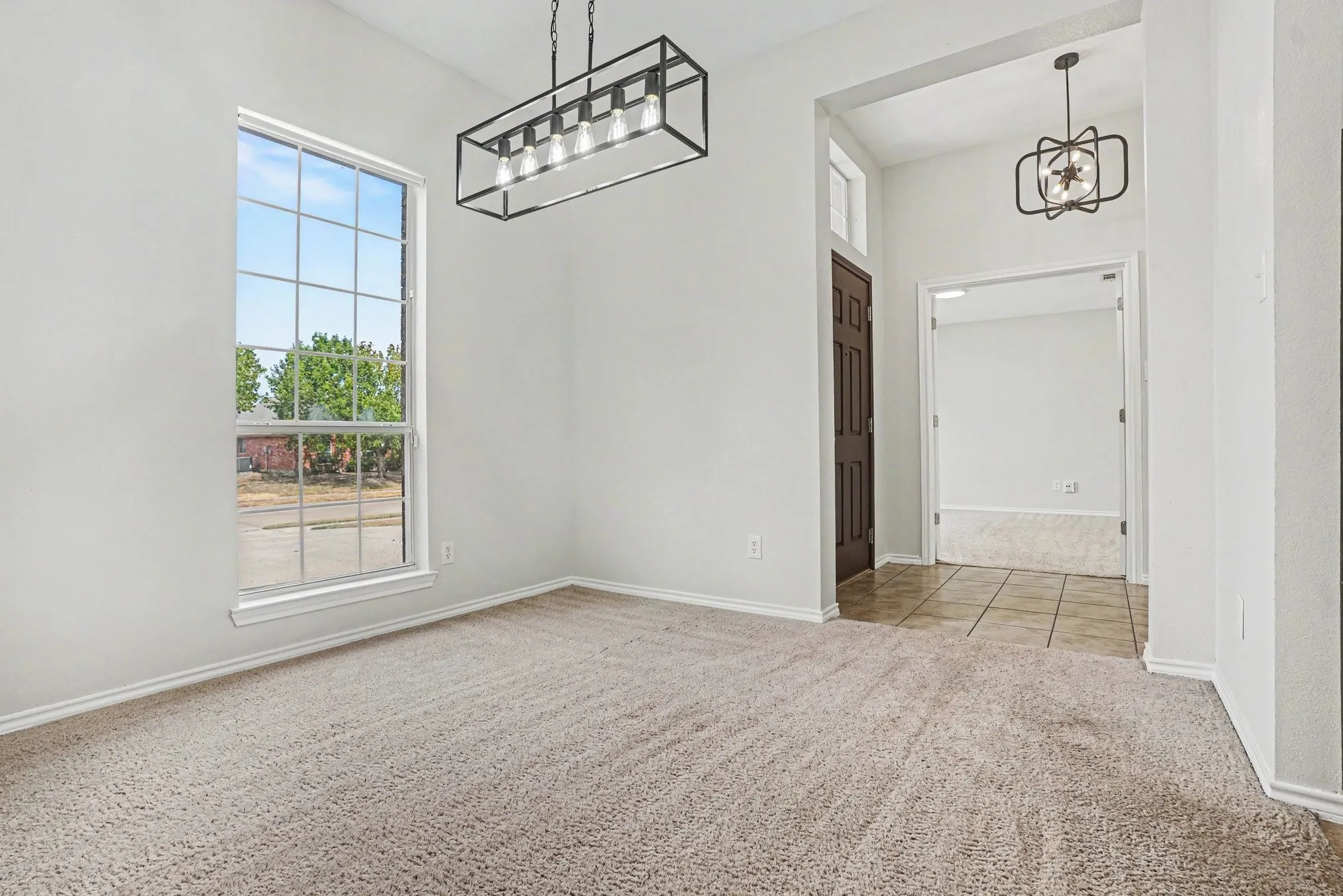 Carpeted room with a large window, a dark wood-paneled door, and a rectangular black metal chandelier