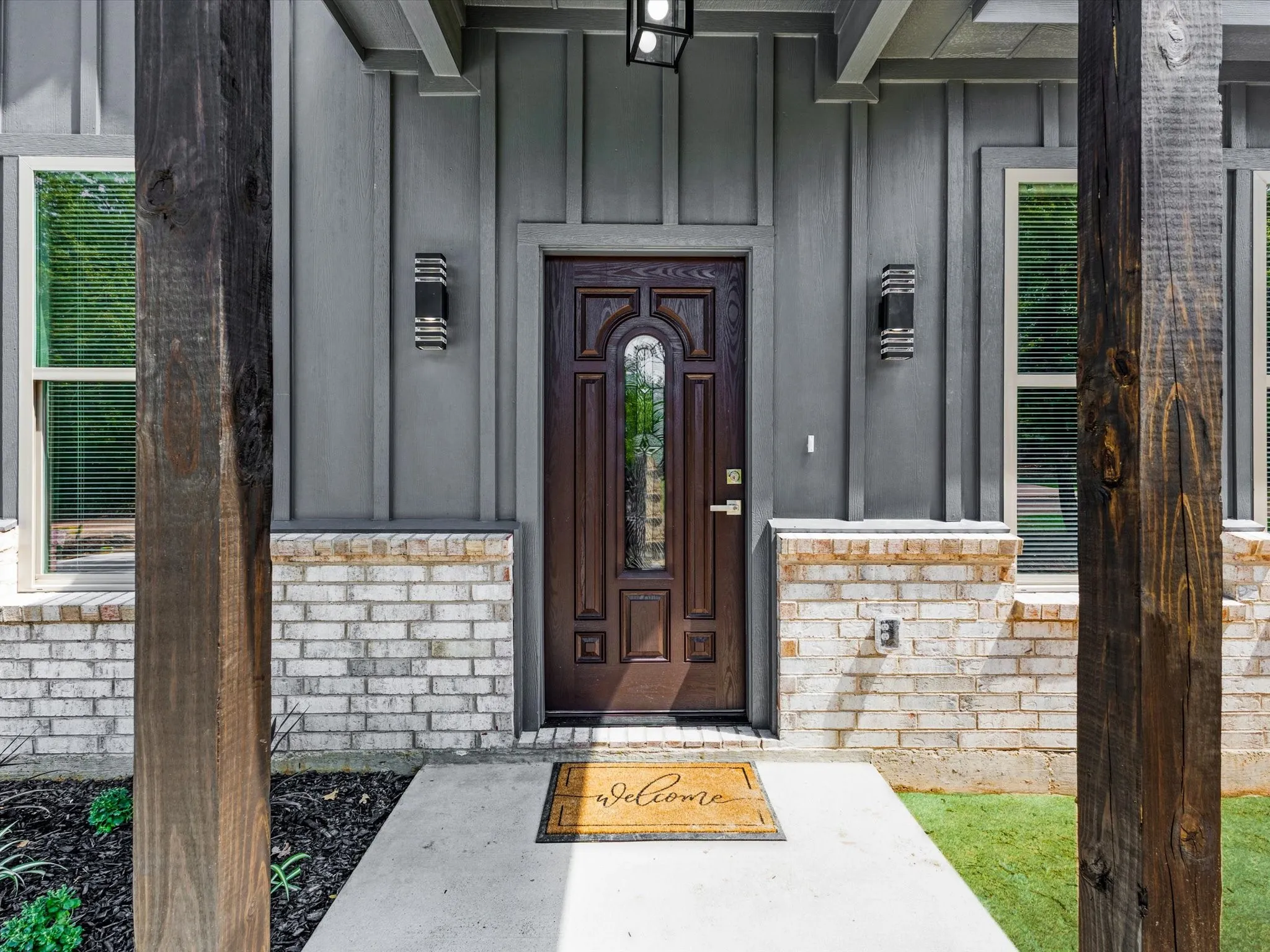 Board and batten siding with a brick facade, wood-finish columns, a dark wood-paneled entry door with glass insert, and modern exterior sconces
