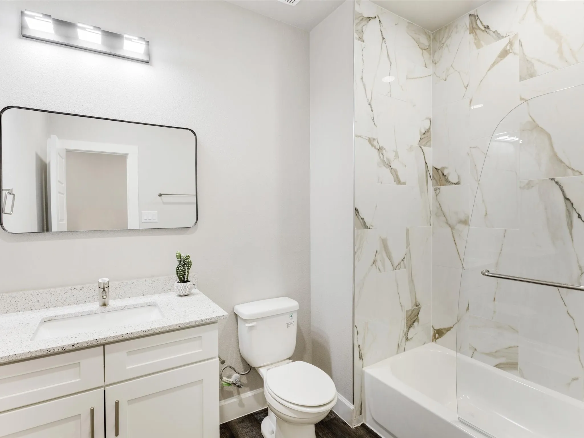 White vanity with a speckled countertop and integrated sink, framed mirror, and a tub-shower combination with floor-to-ceiling veined tile surround