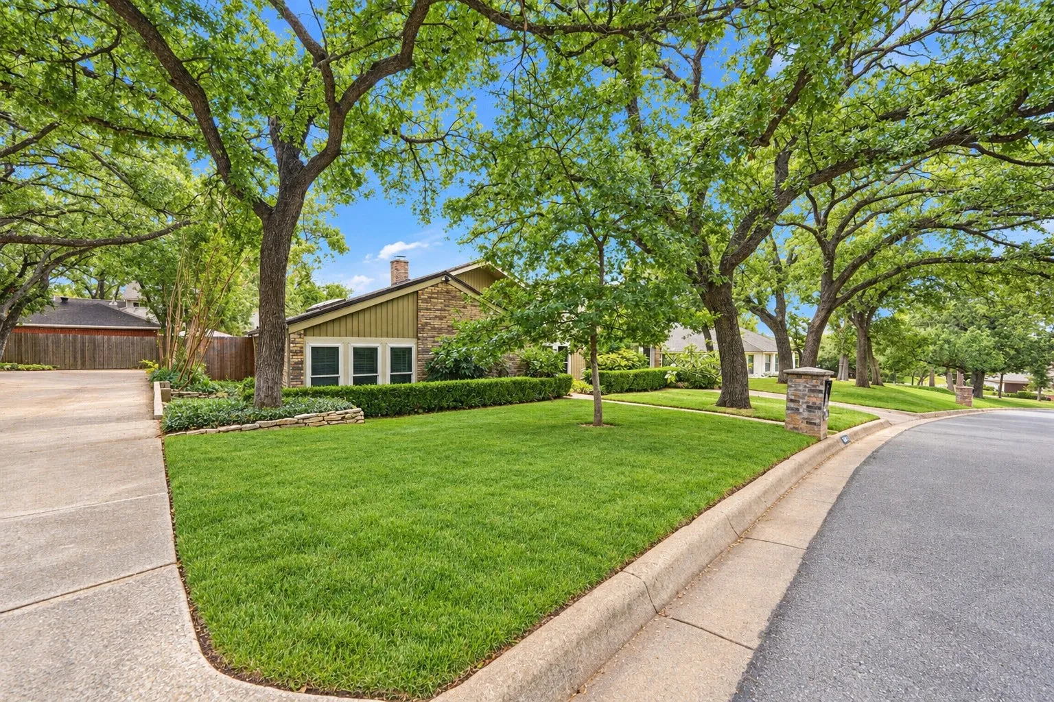 Mature trees, extensive landscaping, stone-clad chimney, manicured lawn, and a paved driveway