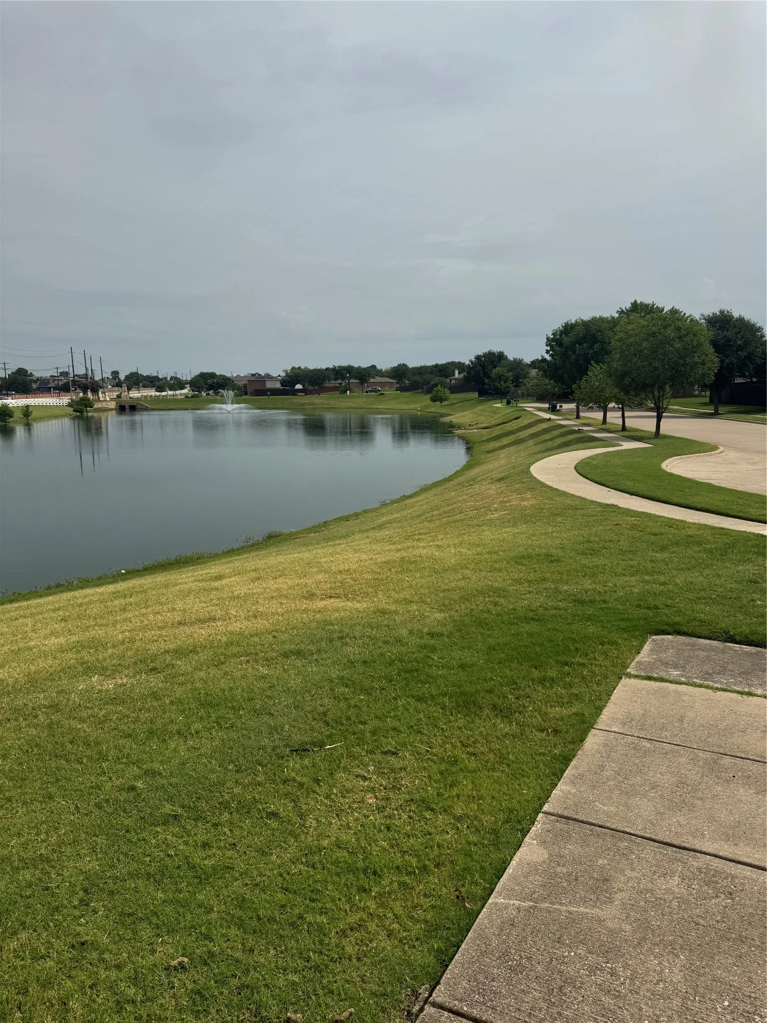 Expansive waterfront with a fountain feature, surrounded by a curving concrete pathway and lush green landscaping
