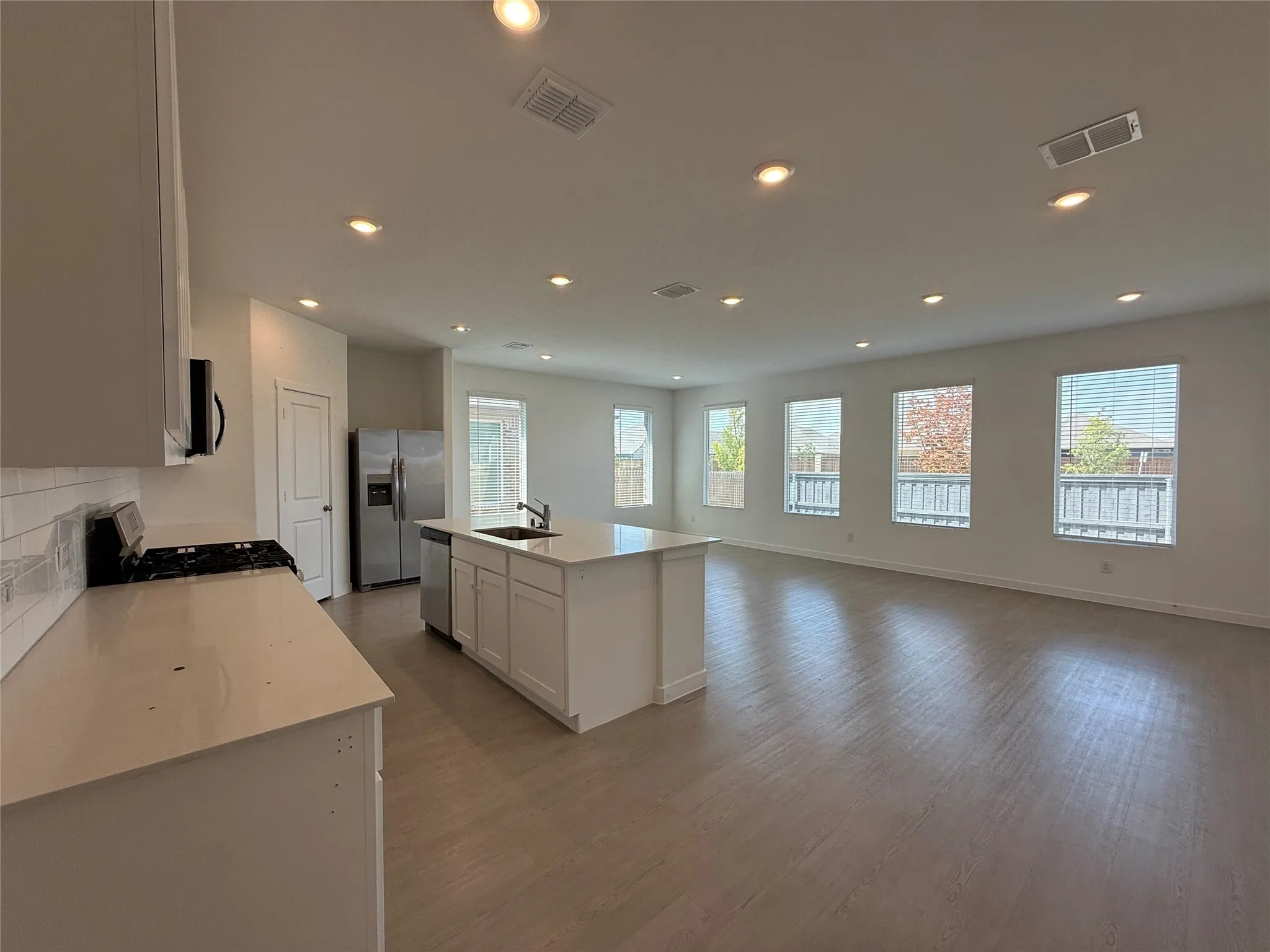 Open-concept living space with recessed lighting, wood-finish flooring, a kitchen island with an integrated sink, white shaker cabinetry, and stainless steel appliances