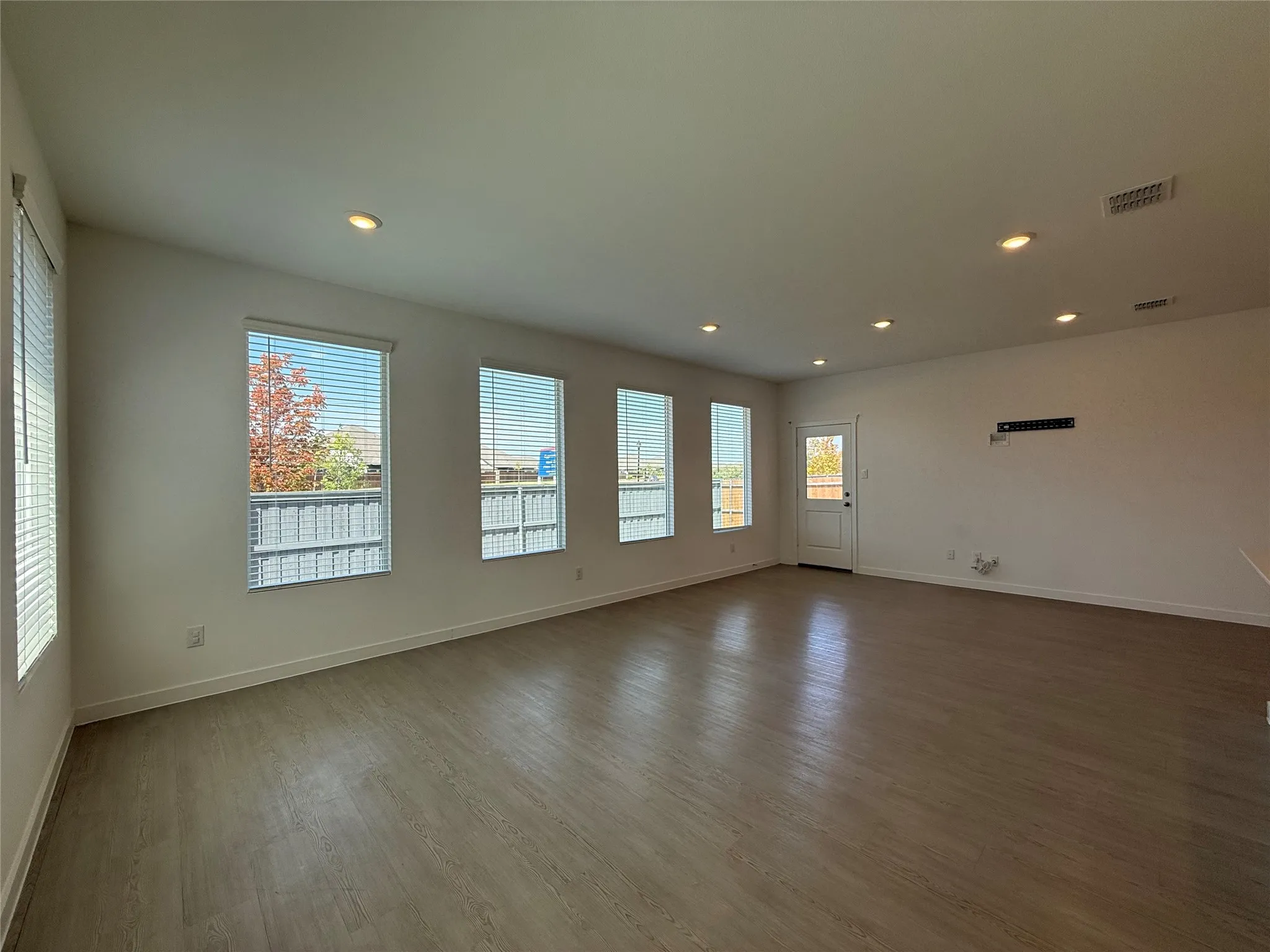 Spacious room featuring wood-finish flooring, recessed lighting, multiple windows with blinds, and a rear door with a full-lite glass insert