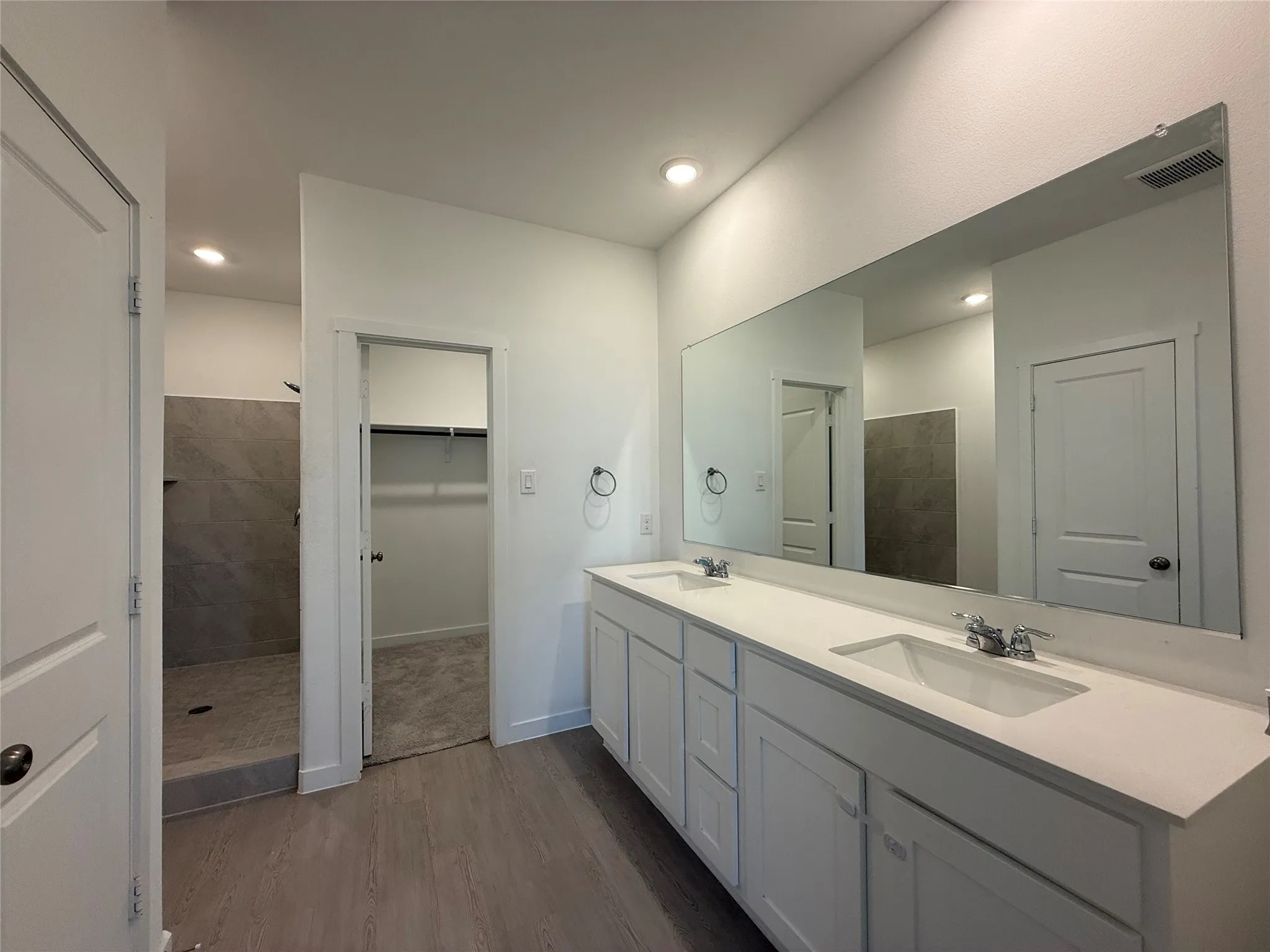 Dual vanity with white cabinetry and integrated sinks, large wall-mounted mirror, wood-finish flooring, and a walk-in shower with tiled walls