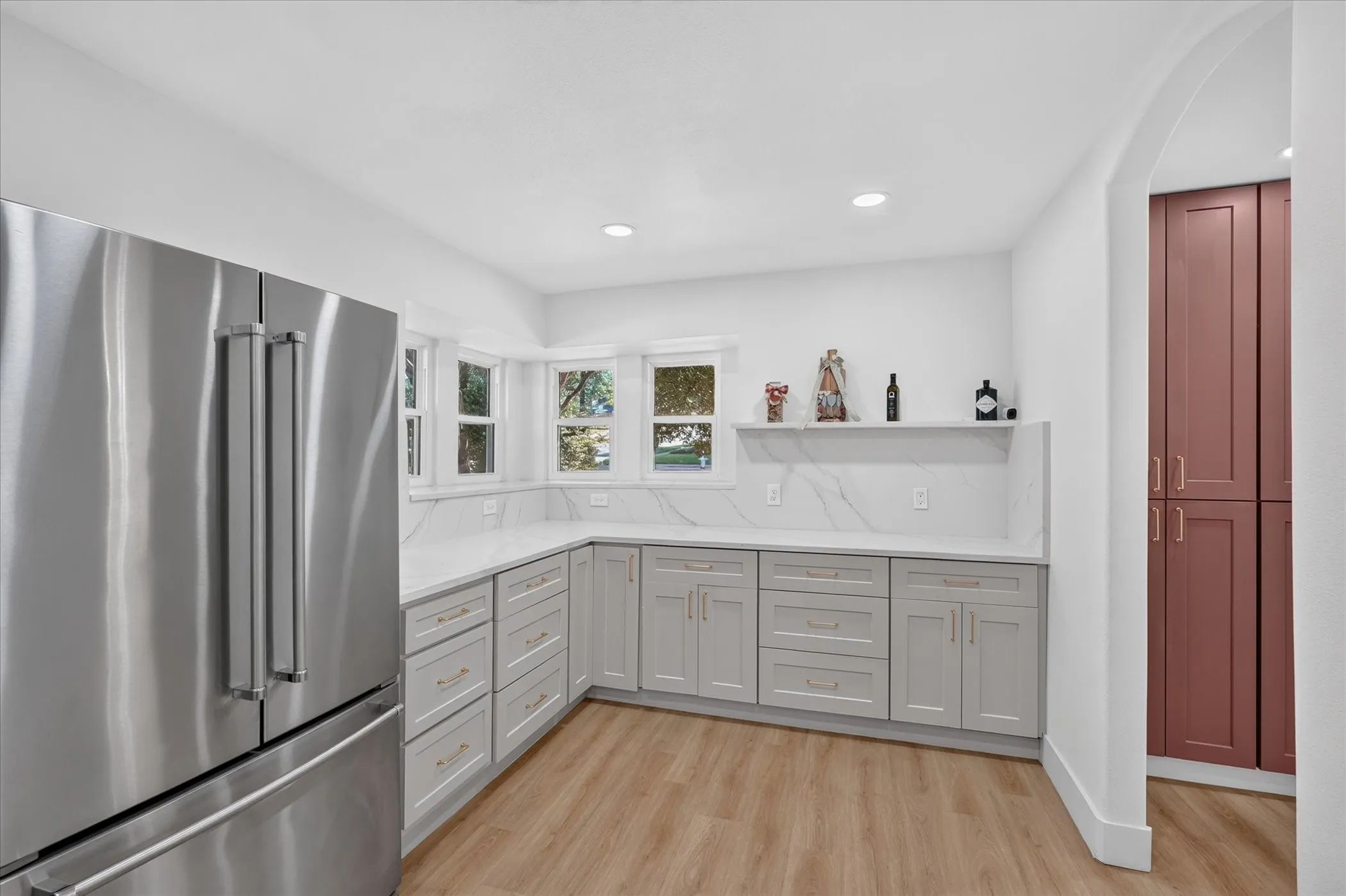 Stainless steel refrigerator, light wood-finish flooring, light gray cabinetry with gold hardware, white countertop with integrated backsplash, and a built-in archway