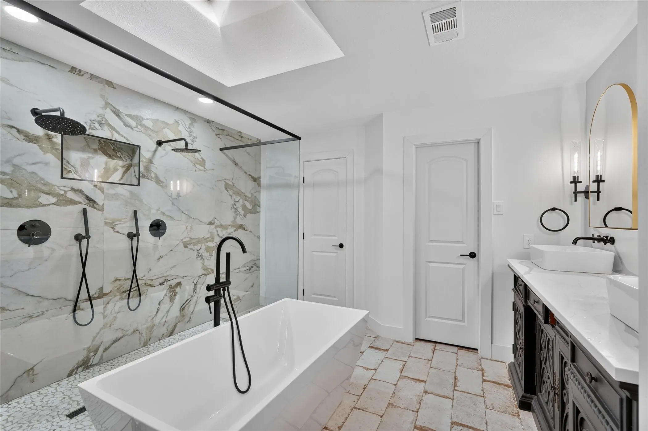 Freestanding soaking tub, walk-in shower with dual shower heads, skylight, double vanity with vessel sinks, and stone-finish floor tiles