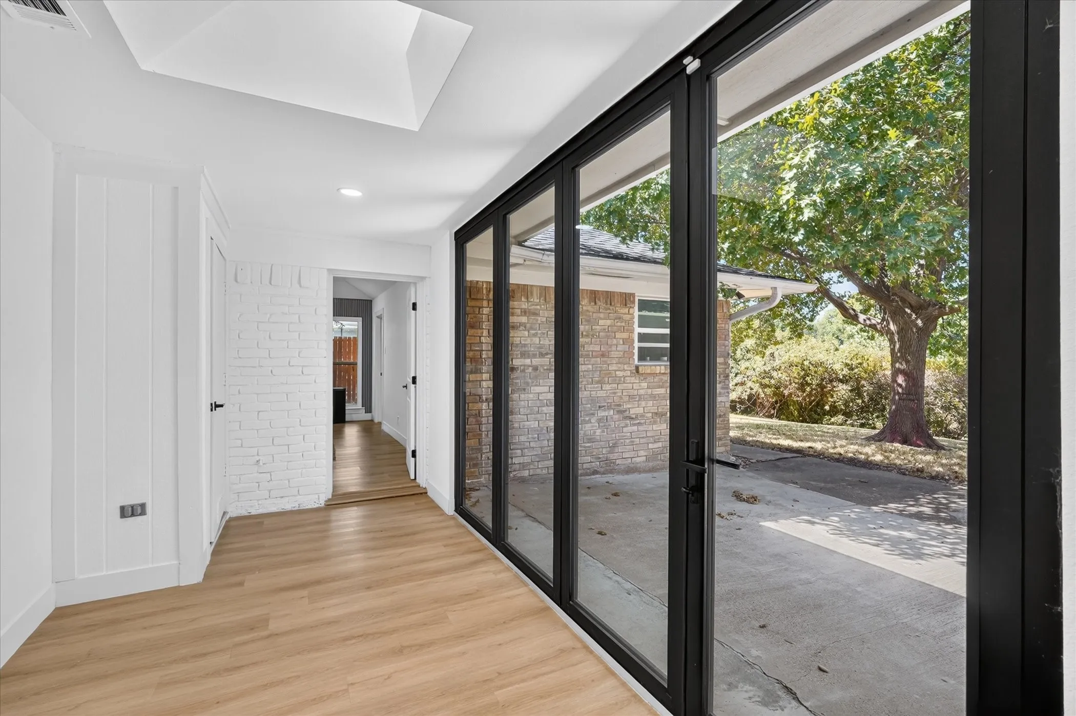 Wood-finish flooring, white shiplap wall, white painted brick wall, black-framed folding glass doors, and a skylight