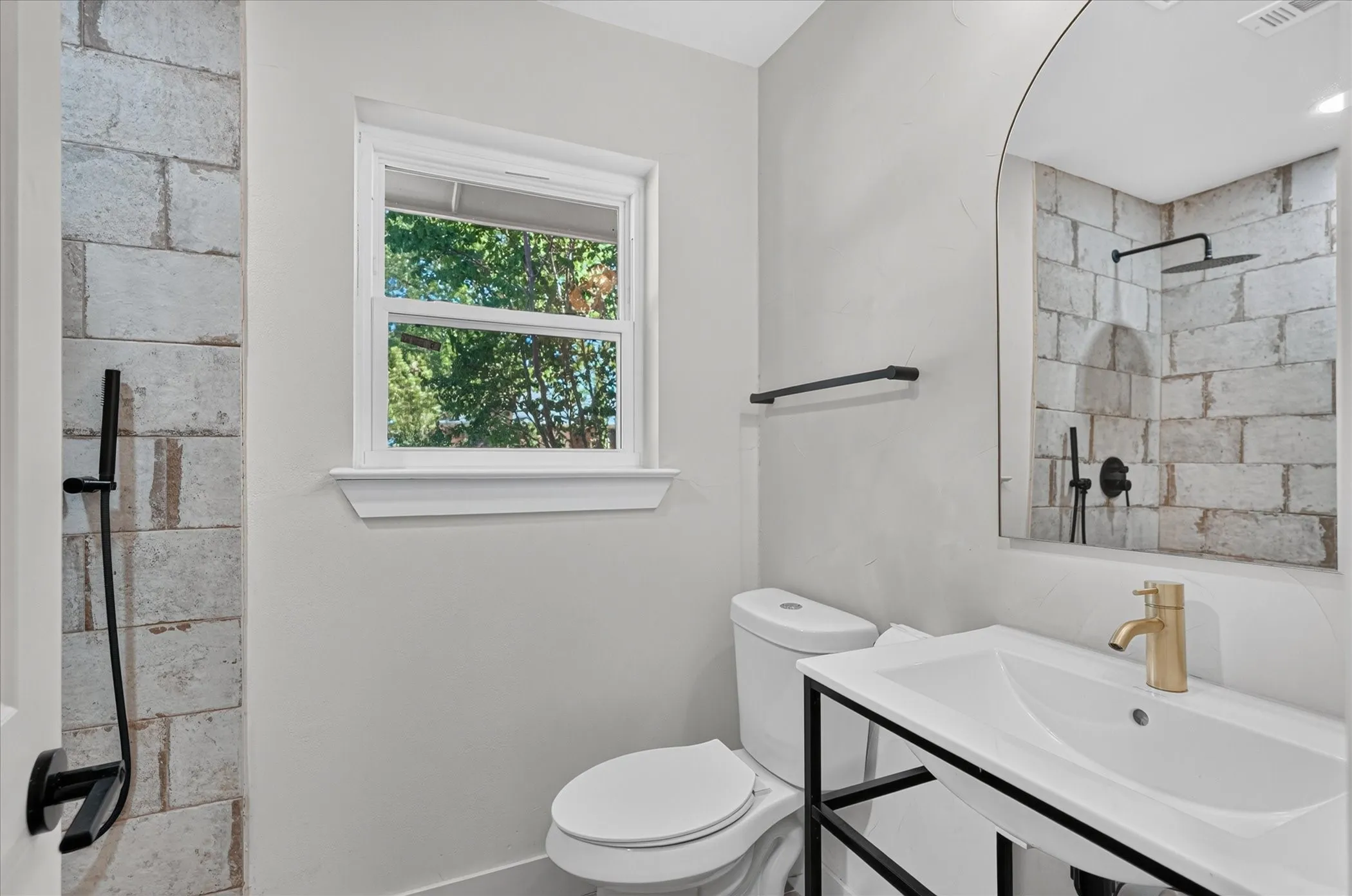 Bathroom featuring a shower with brick-style tiling, a window, a toilet, a vanity with a white basin sink, and an arched mirror