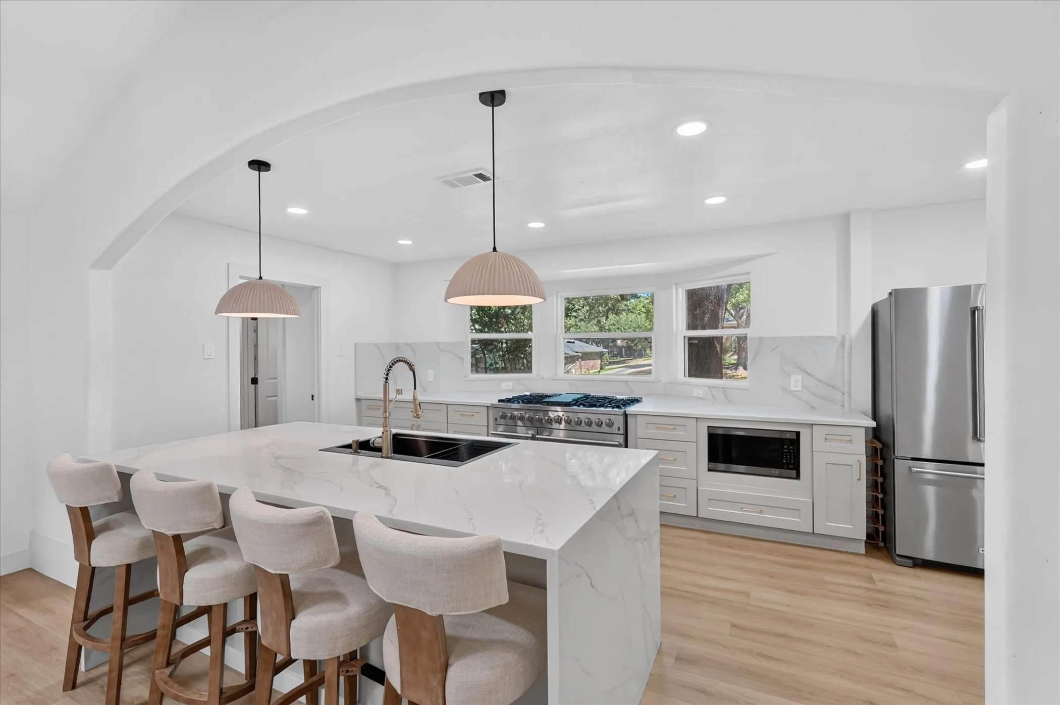 Kitchen featuring a large waterfall island with integrated sink, light wood-finish flooring, stainless steel appliances, light gray cabinetry, and recessed lighting