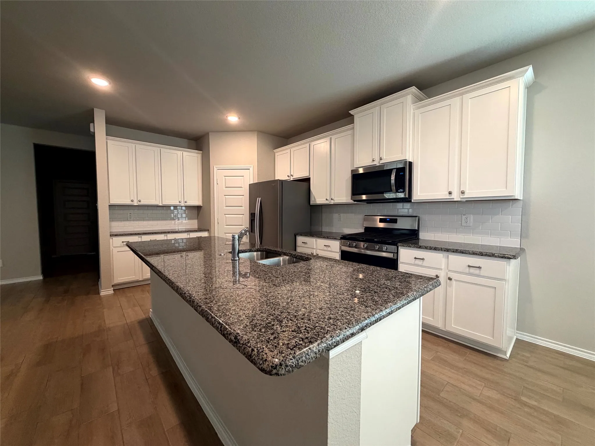 Kitchen with wood-finish flooring, white cabinetry, granite countertops, subway tile backsplash, and stainless steel appliances