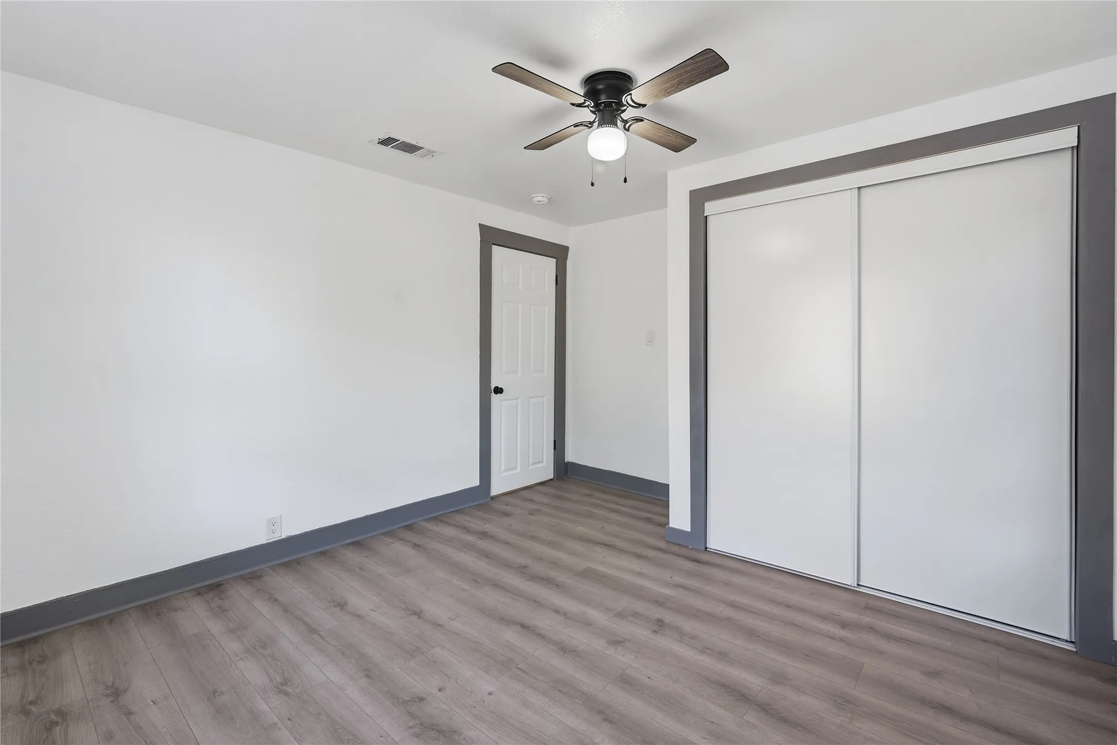 Wood-finish flooring, sliding closet doors, ceiling fan with integrated lighting, white interior door with dark hardware, and contrasting trim