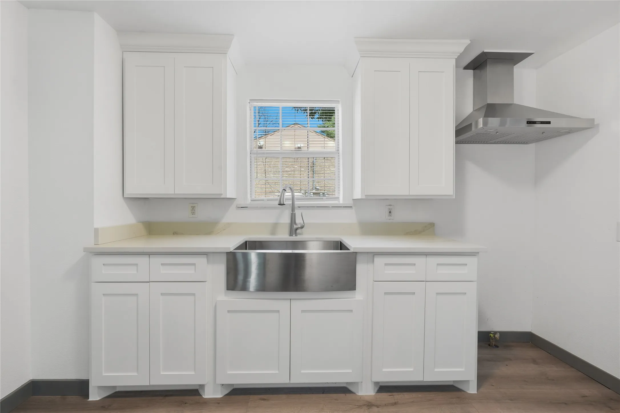 White shaker cabinetry, light-toned countertops, stainless steel farmhouse sink, modern stainless steel range hood, and wood-finish flooring