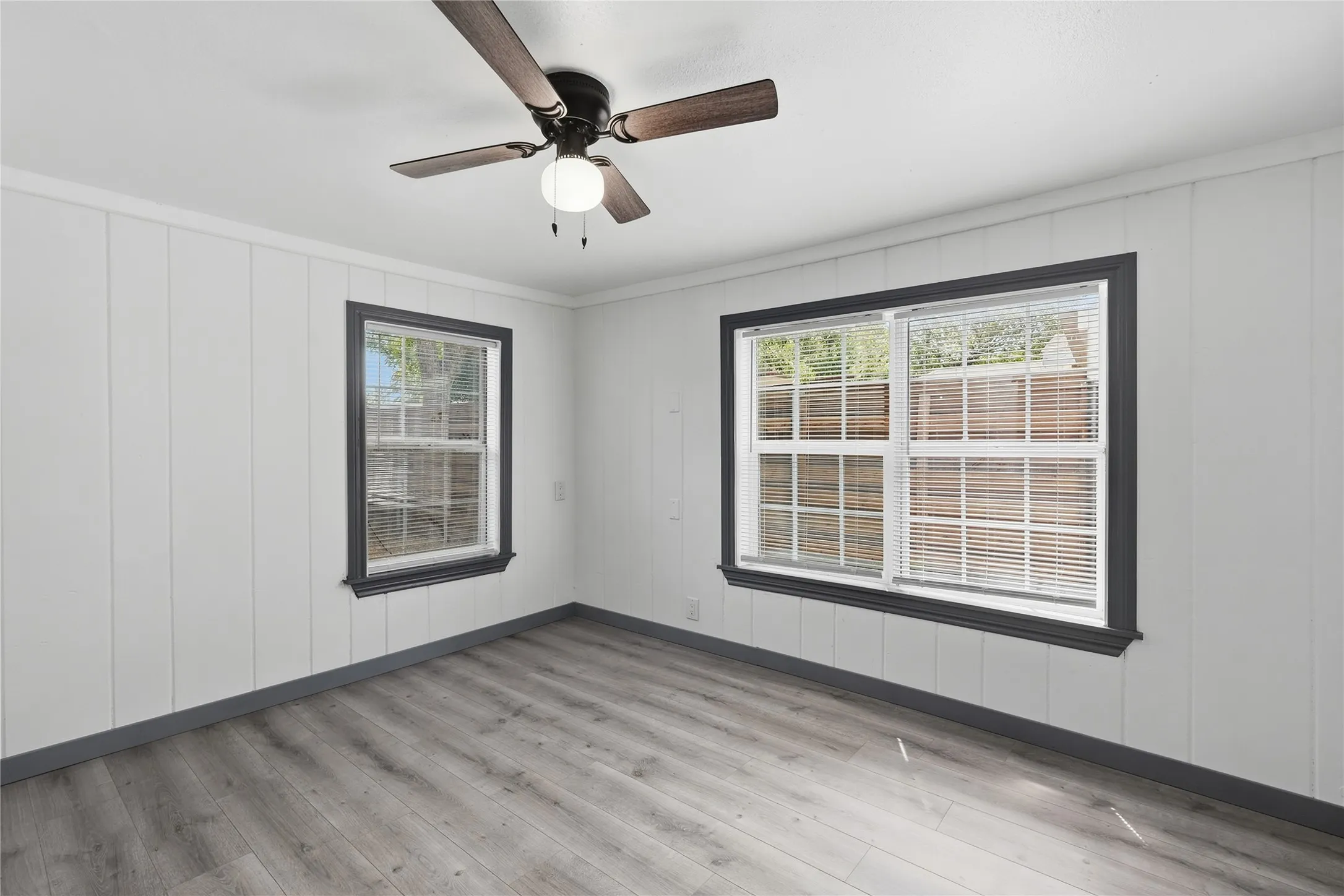 Wood-finish flooring, white paneled walls, ceiling fan with integrated lighting, and two windows with dark trim