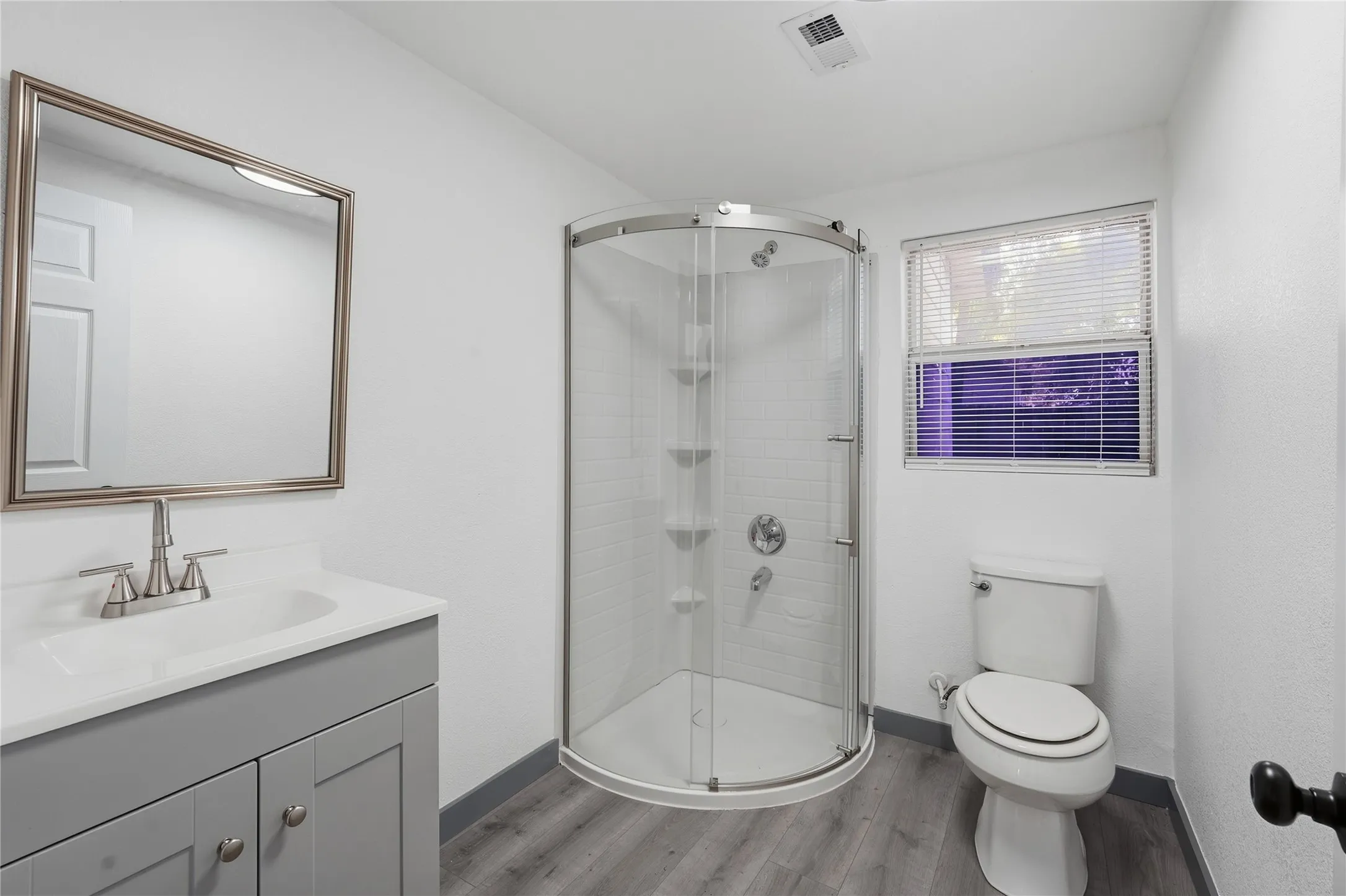 Bathroom featuring a rounded glass-enclosed shower, a vanity with a light gray cabinet and white countertop, a mirror with a brushed nickel frame, a white toilet, and wood-finish flooring