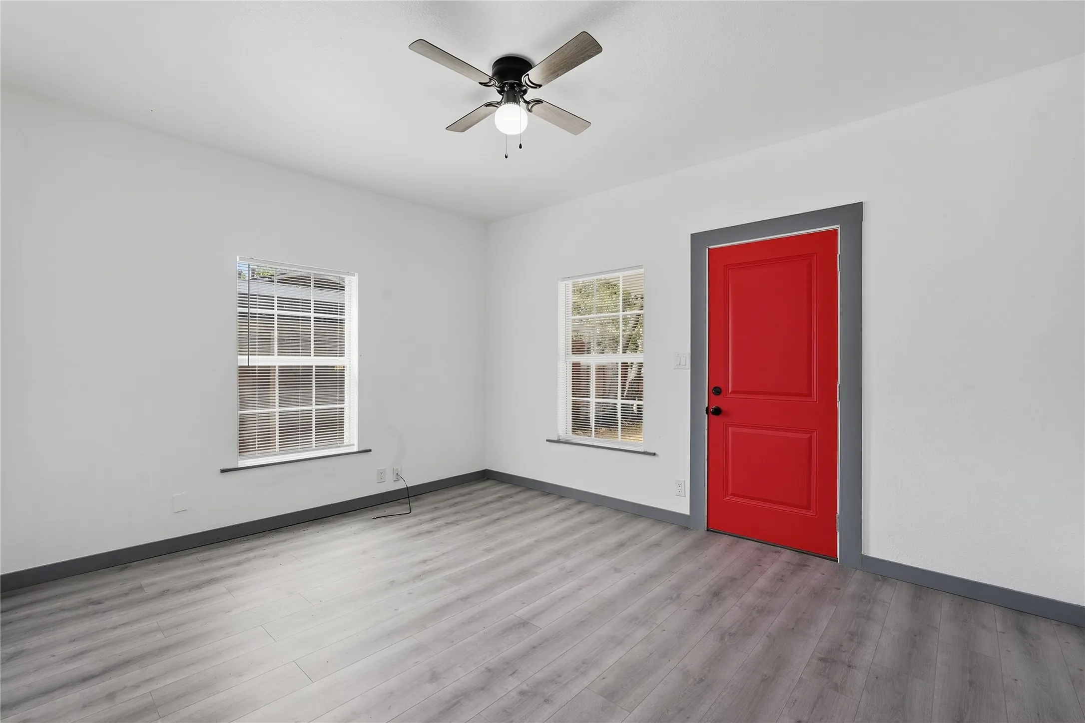 White walls, gray wood-finish flooring, two windows, a red panel door, and a ceiling fan with light fixture