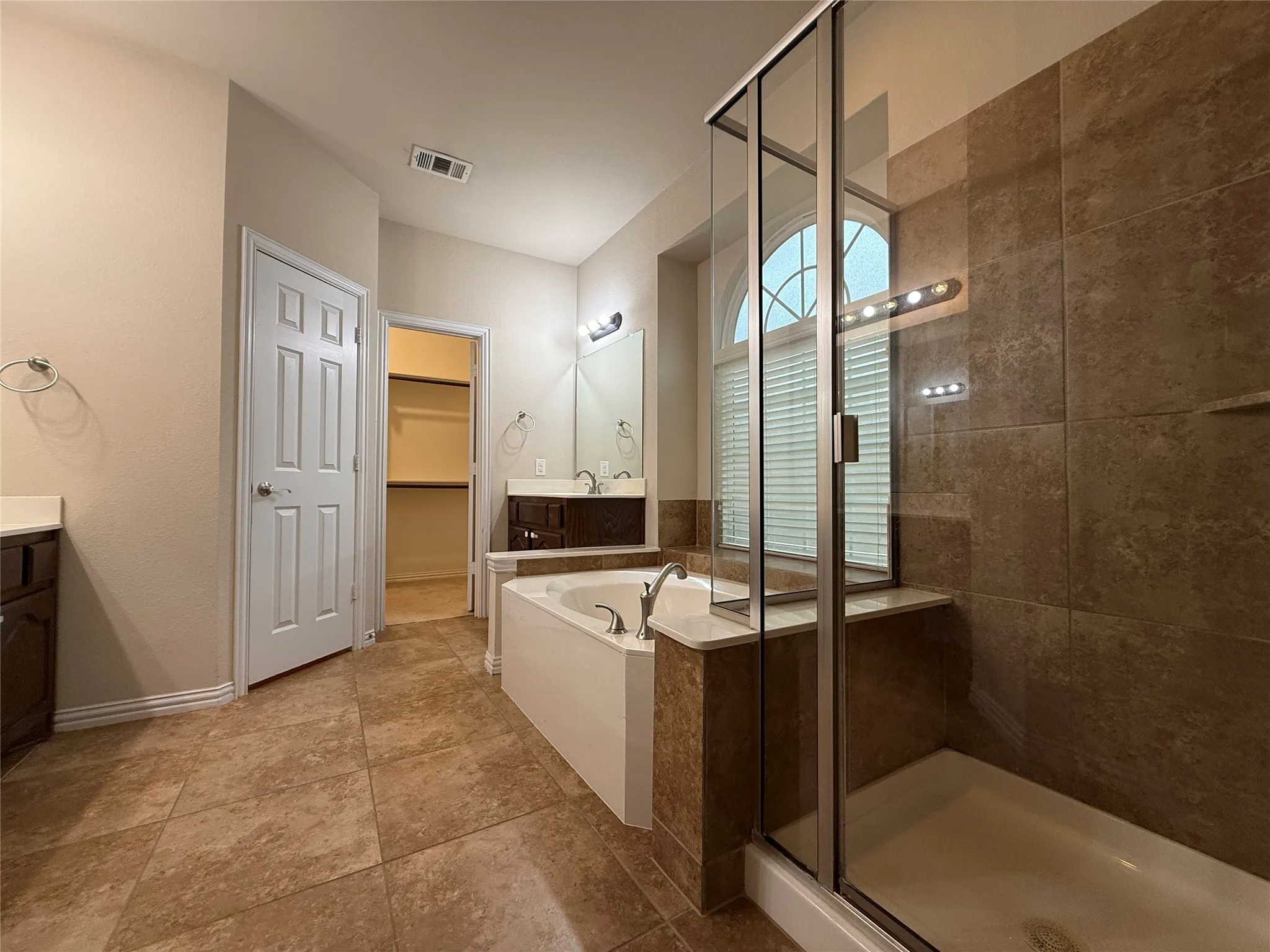 Tiled flooring, glass-enclosed shower, built-in soaking tub, dual vanities with integrated sinks, and an arched window with blinds