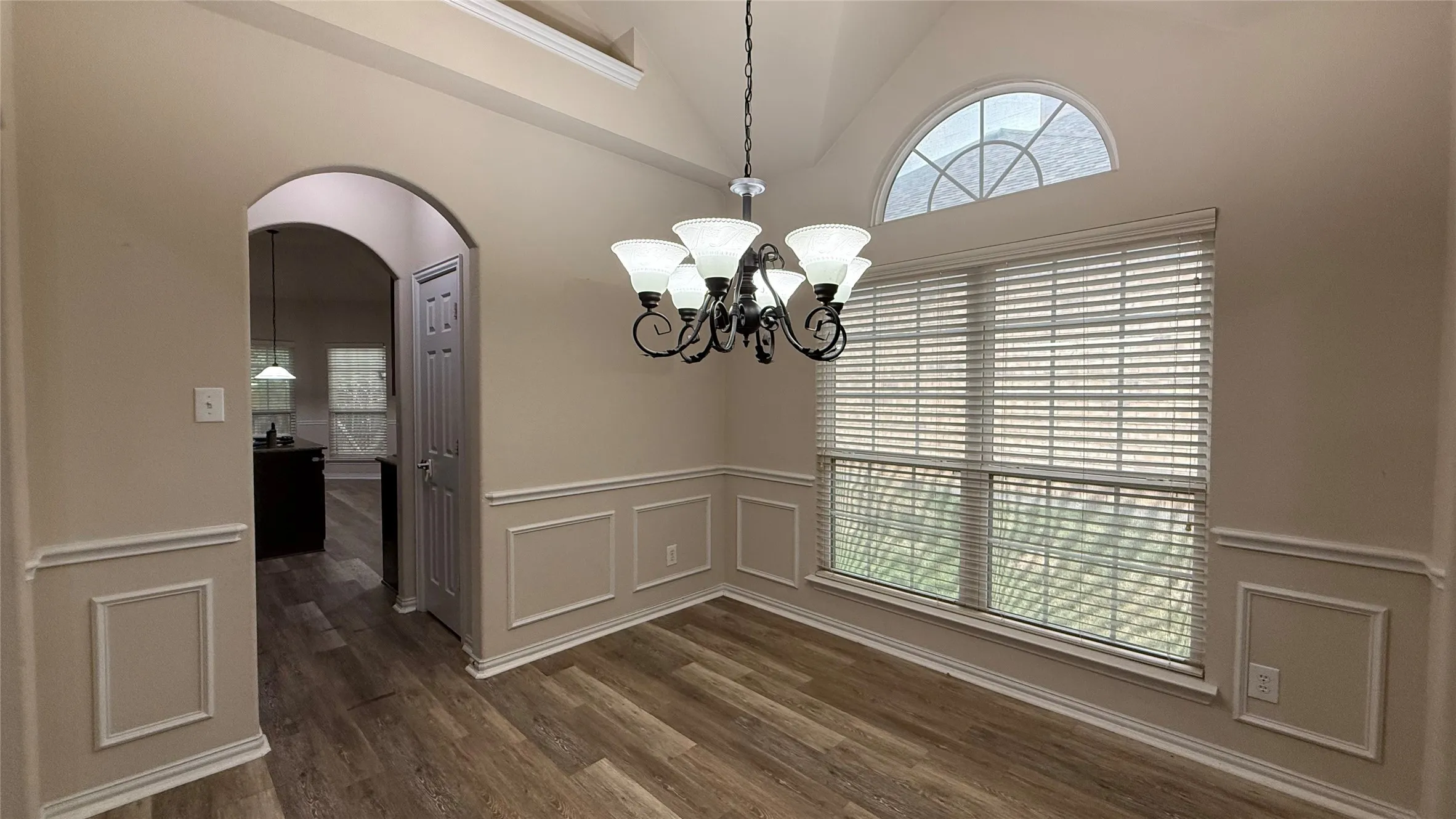 Vaulted ceiling with decorative chandelier, wood-finish flooring, wainscoting, arched doorway, and large window with arched transom