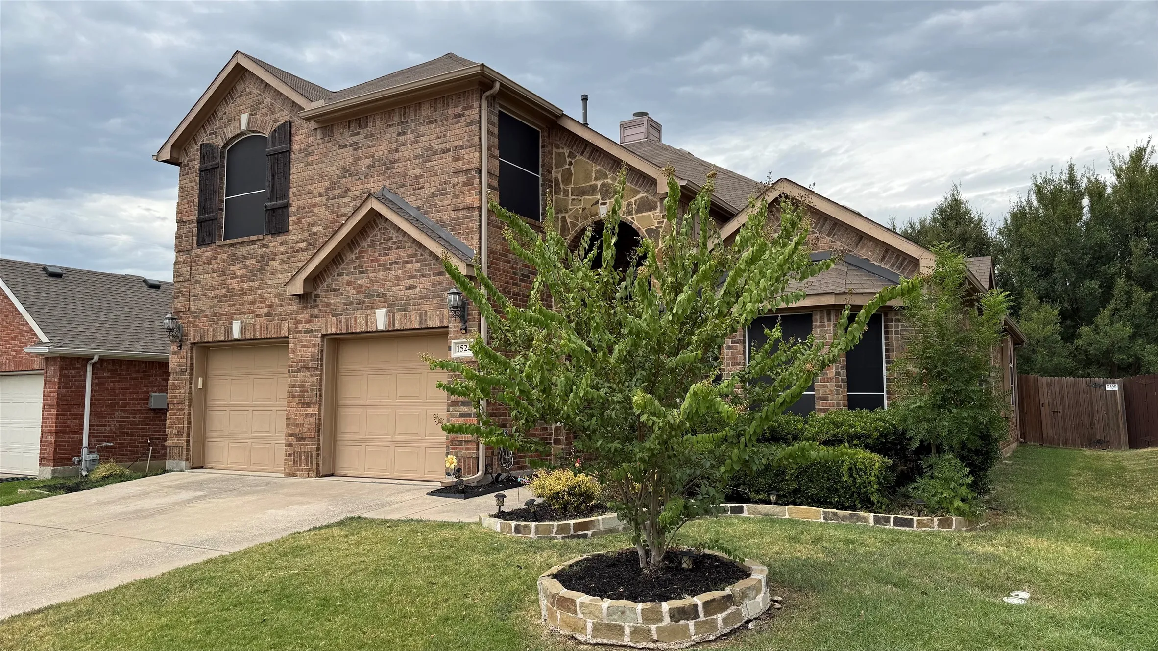Brick and stone façade with a two-car garage, mature landscaping, and a fenced yard