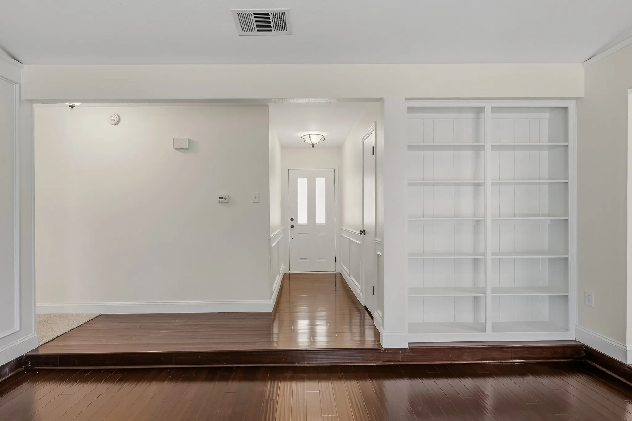 Welcoming entry opens to a spacious living area featuring rich wood-look flooring, crisp neutral finishes, and detailed wall paneling. Custom built-in shelving provides attractive display and storage space, while the raised living room creates a subtle separation between spaces. A wide hallway leads toward the front entry, enhancing the home’s open and inviting feel.