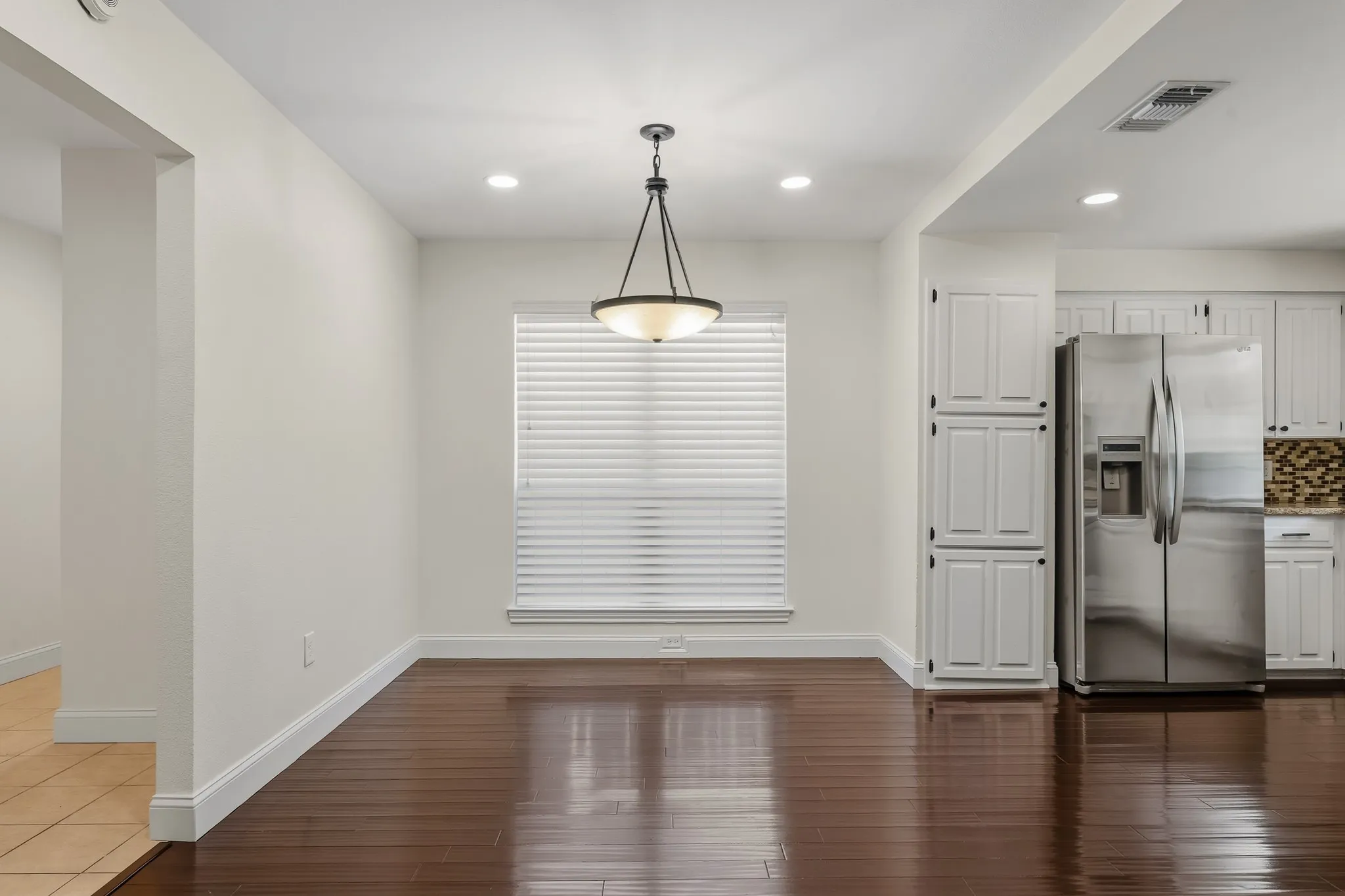 Bright and inviting dining area featuring rich wood-look flooring, neutral finishes, and a stylish pendant light. A large window with blinds provides natural light, while the open layout offers seamless flow into the kitchen. Ample wall space and a comfortable layout make this an ideal setting for everyday dining or entertaining.